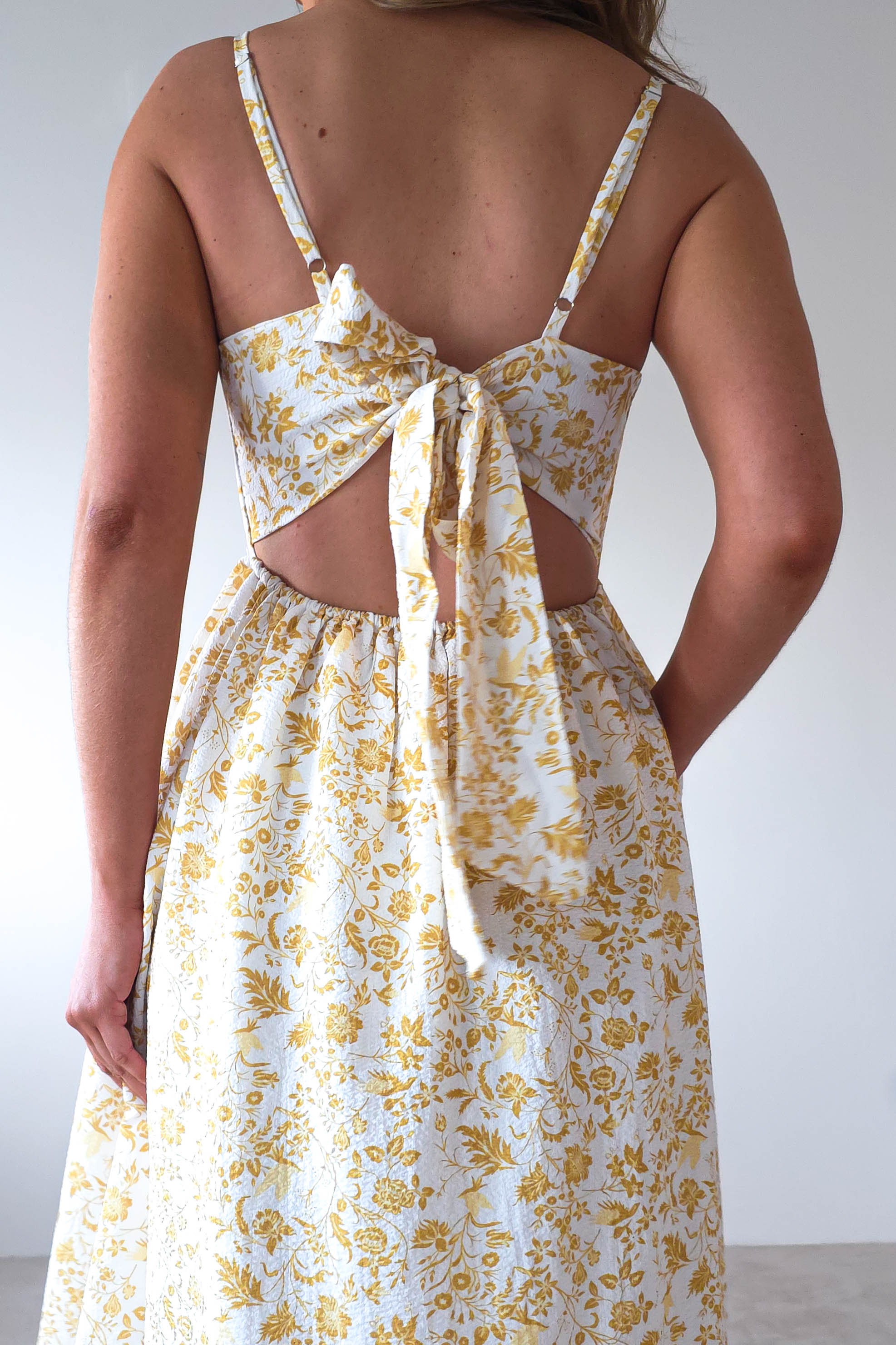 Back view of a person wearing a sleeveless, yellow and white floral summer print maxi dress. The dress features a cut-out on the back with adjustable ties forming a tied bow detail. Perfect for wedding guests, they stand against a plain light-colored background.