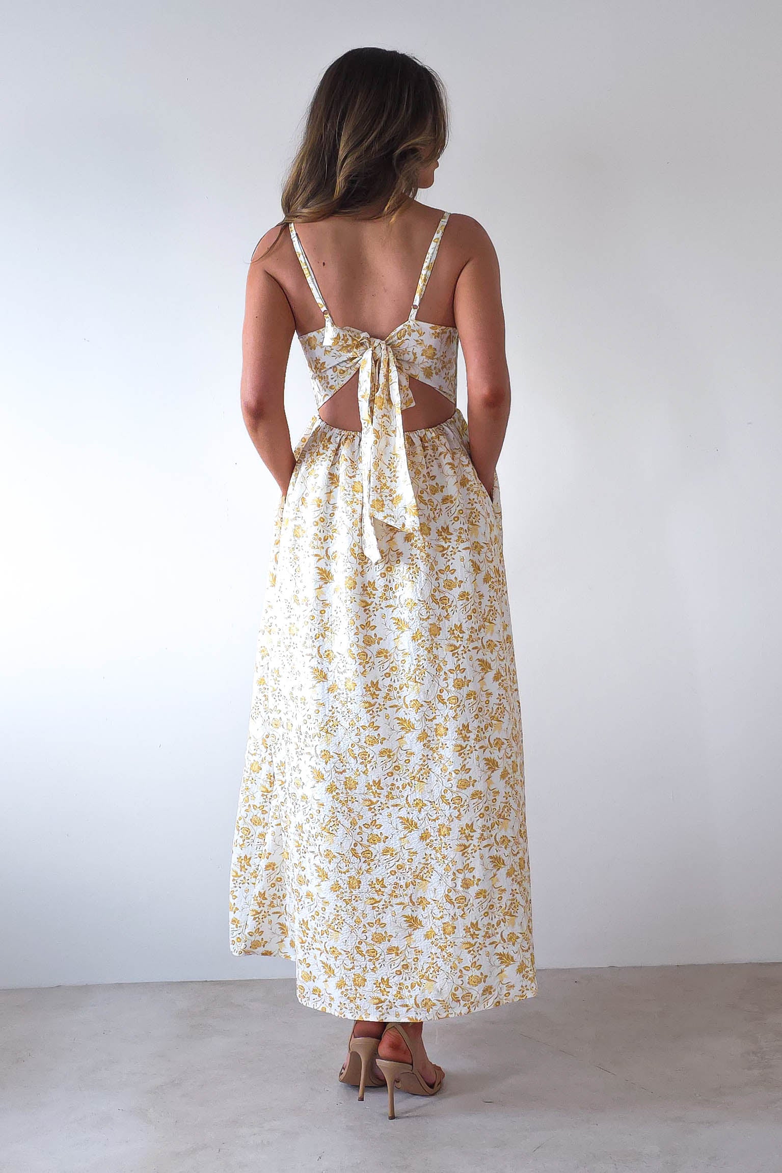 A person with long hair stands poised in a sleeveless summer print maxi dress featuring adjustable tie-back details, facing a white wall. As they wear high heels, their hands rest relaxed at their sides on the light, smooth floor—perfect attire for wedding guests.