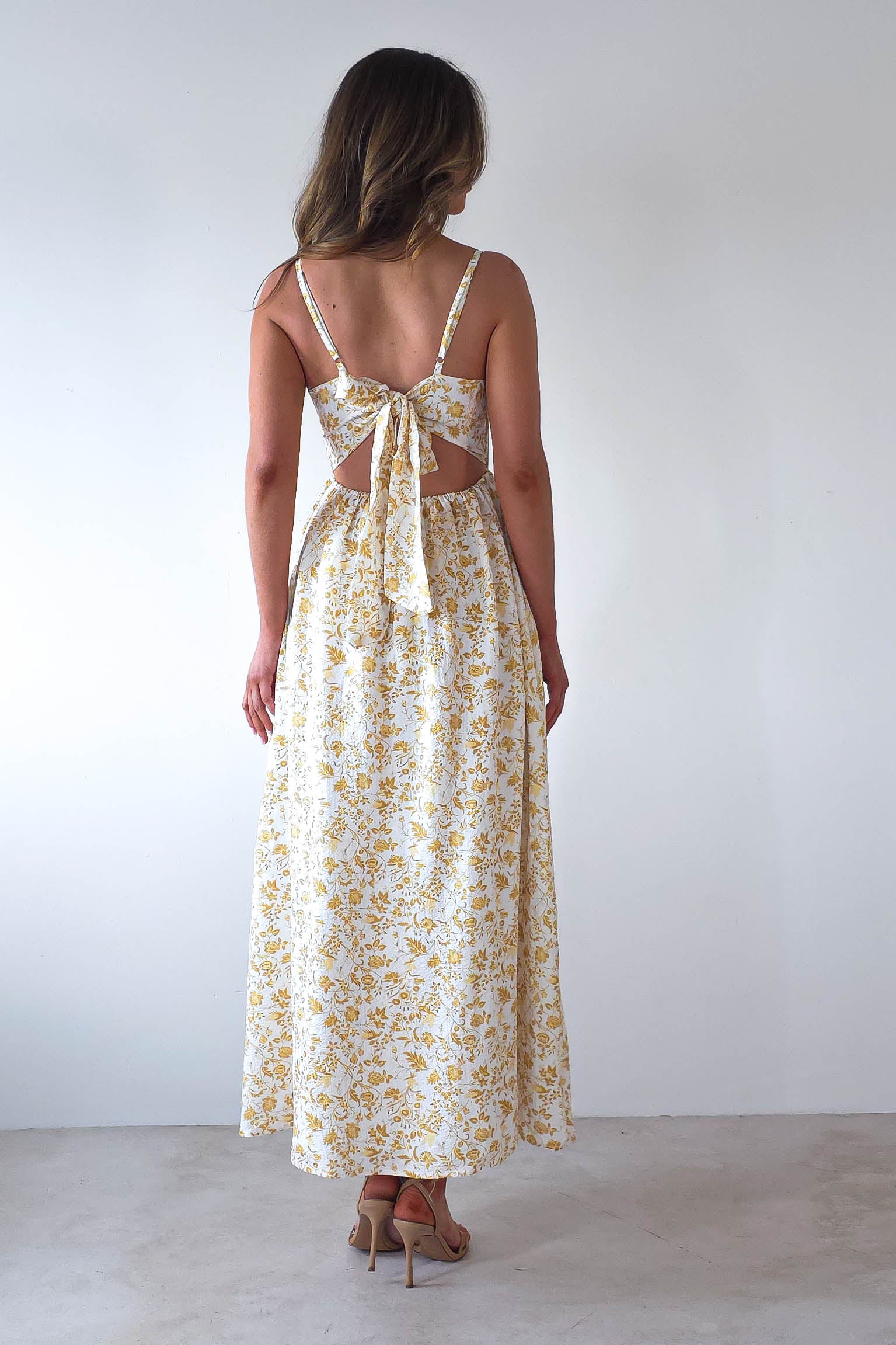 Rear view of a woman wearing a summer print maxi dress with a yellow floral pattern. The dress features thin, adjustable ties as straps, a large bow, and a cut-out detail at the back. She stands against a plain white background and wears beige high heels.