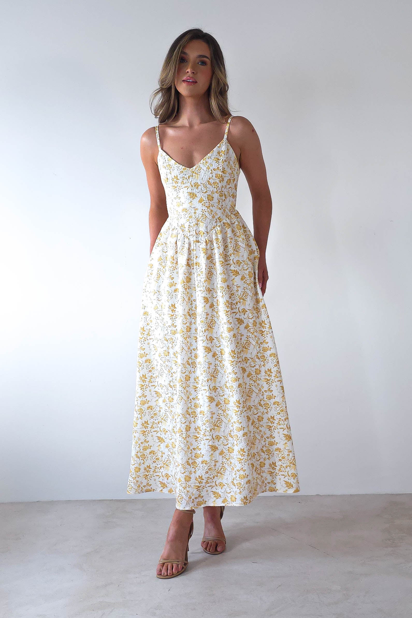 A woman poses against a stark white backdrop, donning a summer print maxi dress adorned with vibrant yellow florals. With long, flowing hair and thin straps with adjustable ties, she exudes effortless grace—a perfect choice for wedding guests seeking style and comfort.