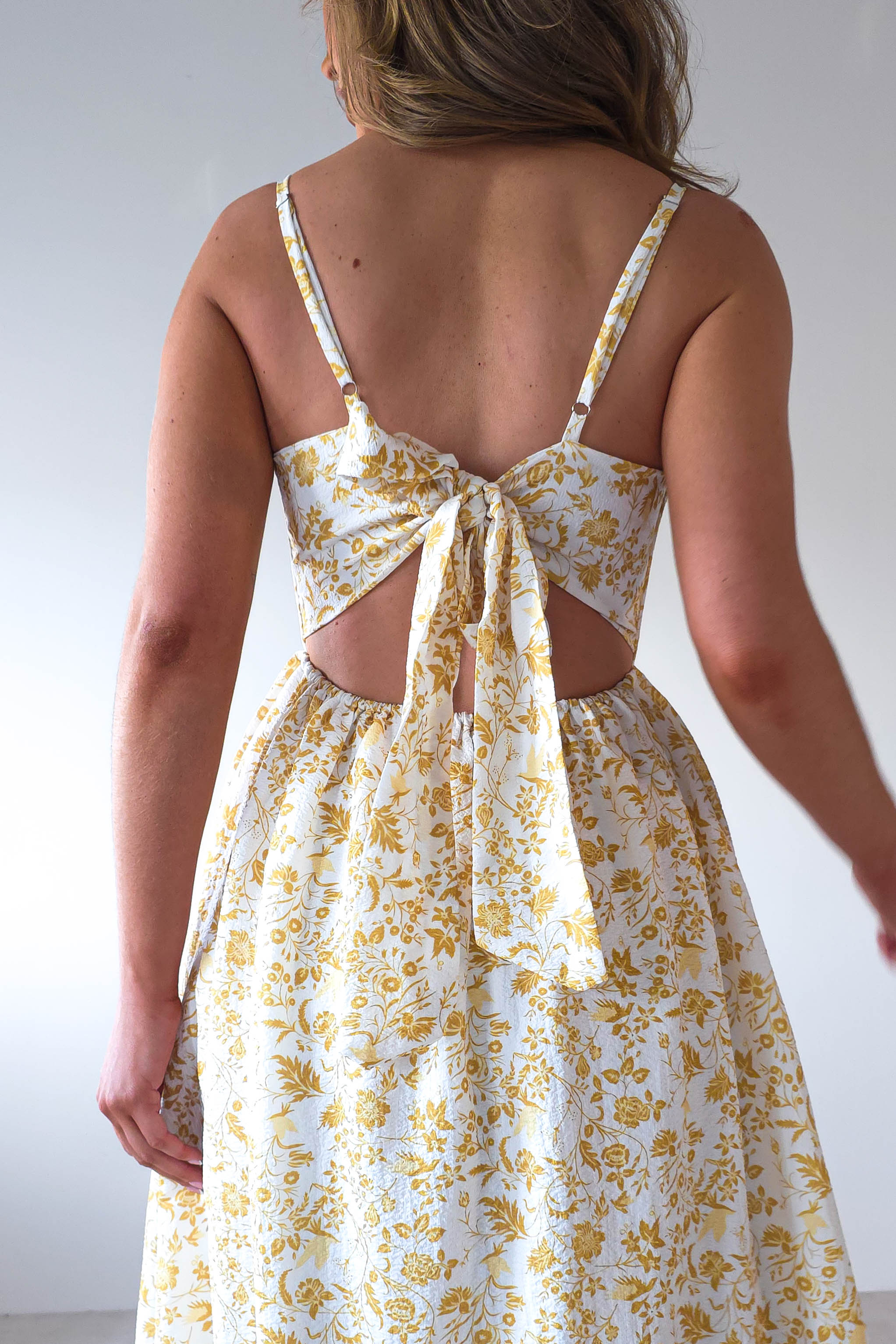 Rear view of a person wearing a sleeveless dress with a yellow floral pattern, perfect for wedding guests. The summer print maxi dress features adjustable ties at the back and a charming cut-out section. The background is a plain white wall.