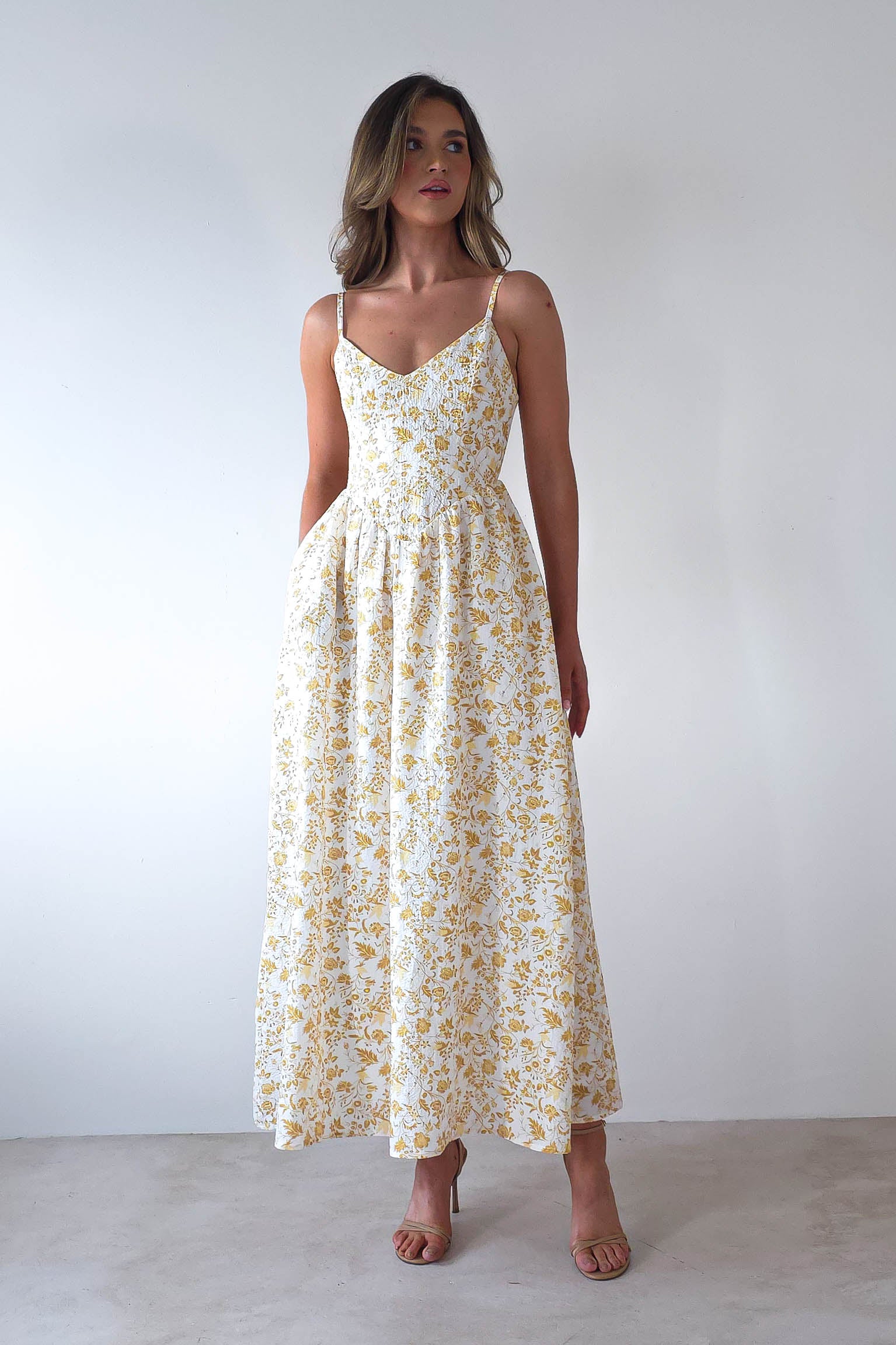 A woman poses against a white backdrop in a summer print maxi dress adorned with yellow floral patterns. The dress features thin straps and a V-neckline, complemented by adjustable ties. She completes her elegant look with beige high heels and wavy hair, perfect for wedding guests.