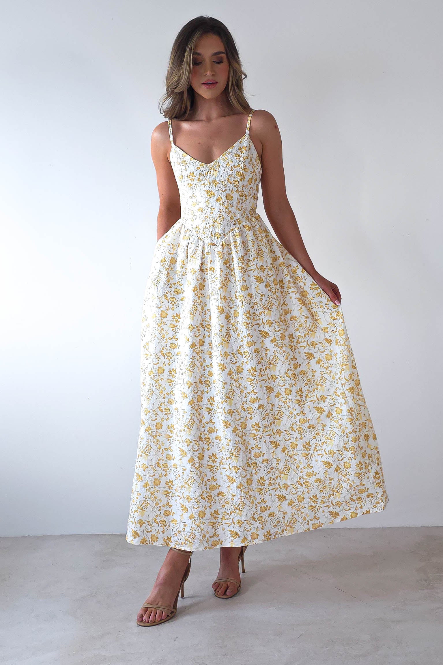 A woman stands against a plain white background wearing a sleeveless, summer print maxi dress with a yellow floral pattern. She holds the sides of the dress, and is wearing strappy heels, with her hair styled loosely—a perfect choice for wedding guests.