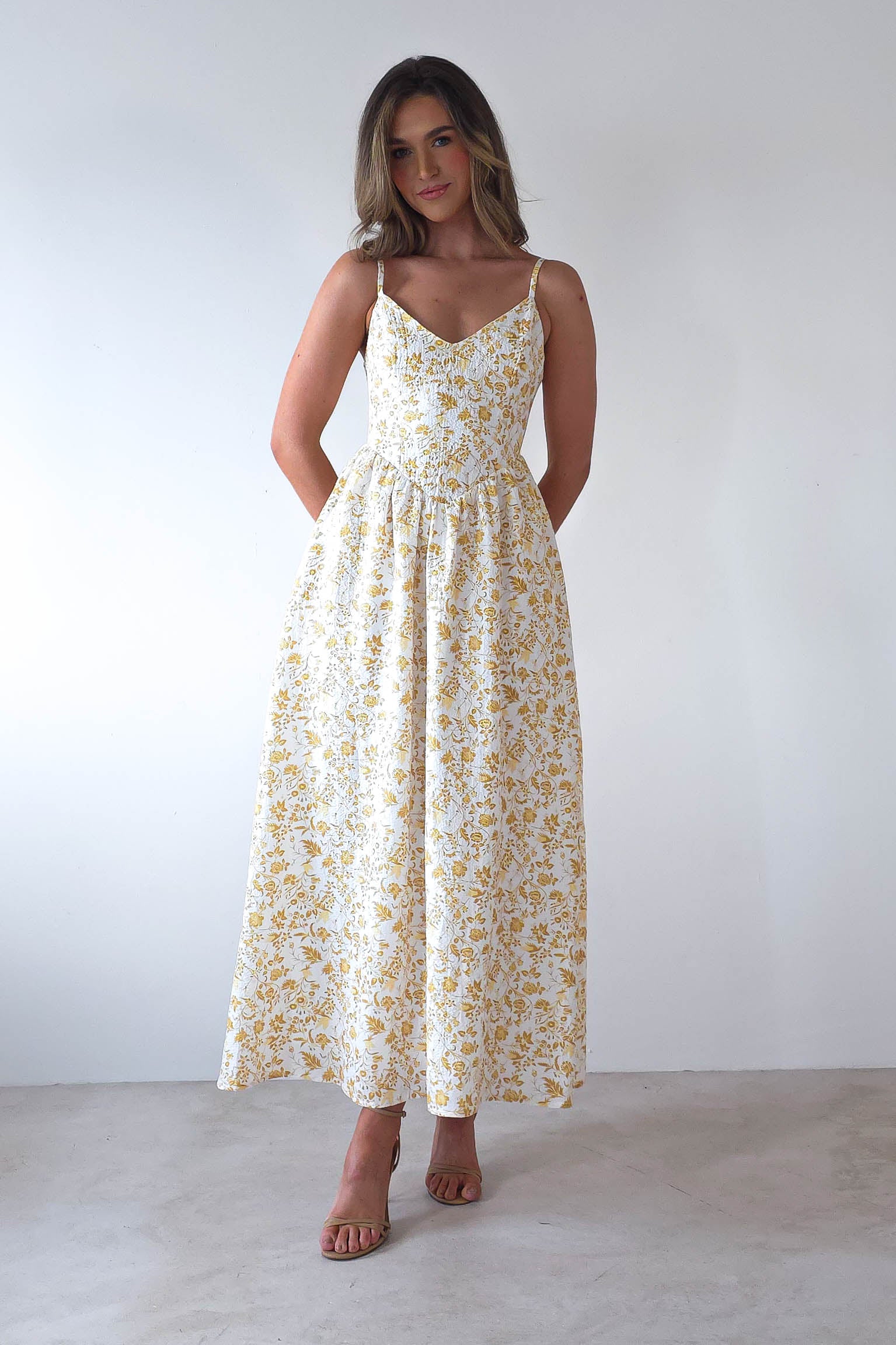 The woman stands elegantly, showcasing a summer print maxi dress adorned with yellow florals. With adjustable ties and brown sandals complementing her look, she exudes an air of grace suitable for wedding guests. Her hair cascades freely against the plain white backdrop.