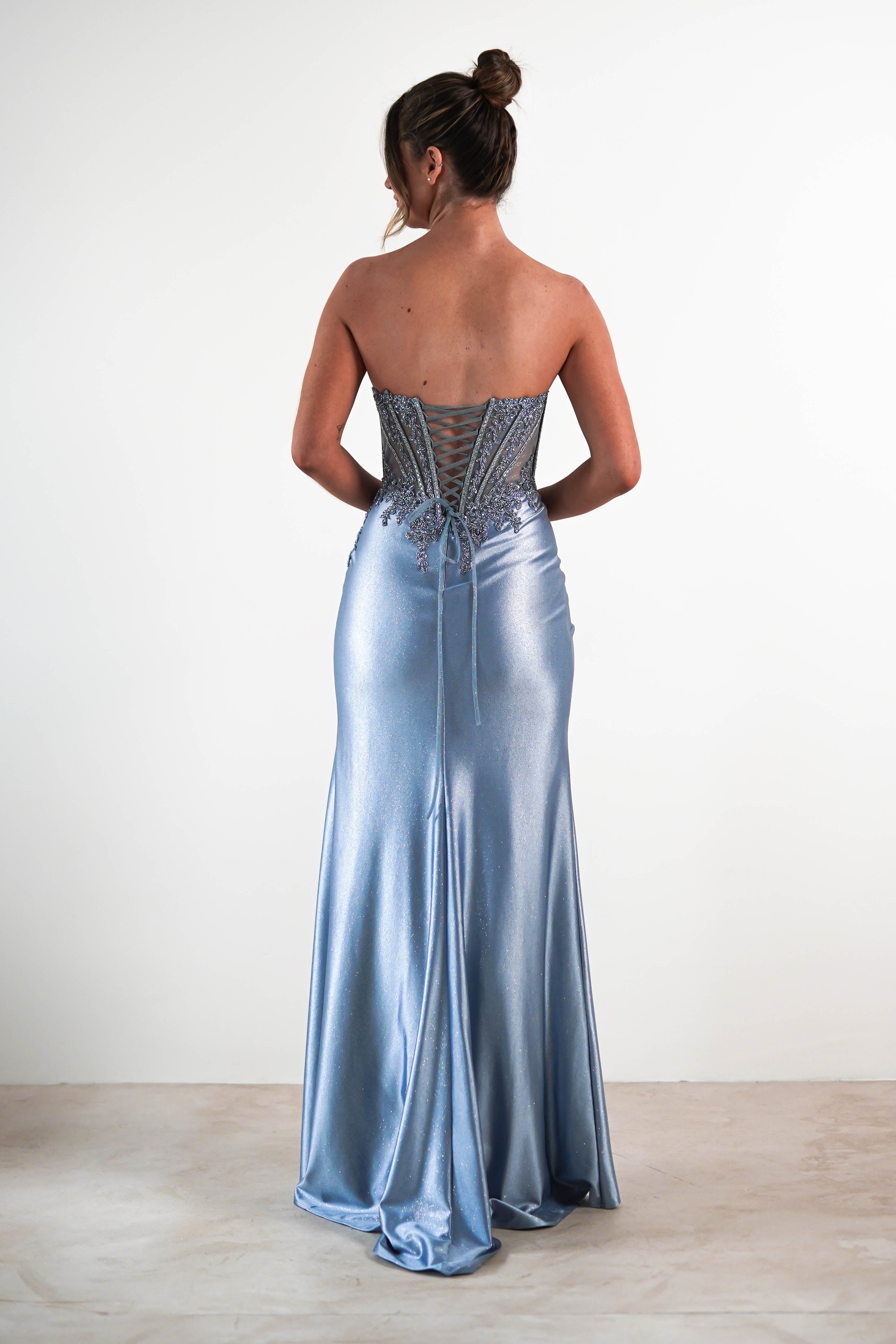 A woman wearing a strapless, sequin embellished gown with a lace-up corset back stands facing away from the camera, her hair in a neat bun. The dress features a fitted bodice and a long, flowing skirt.