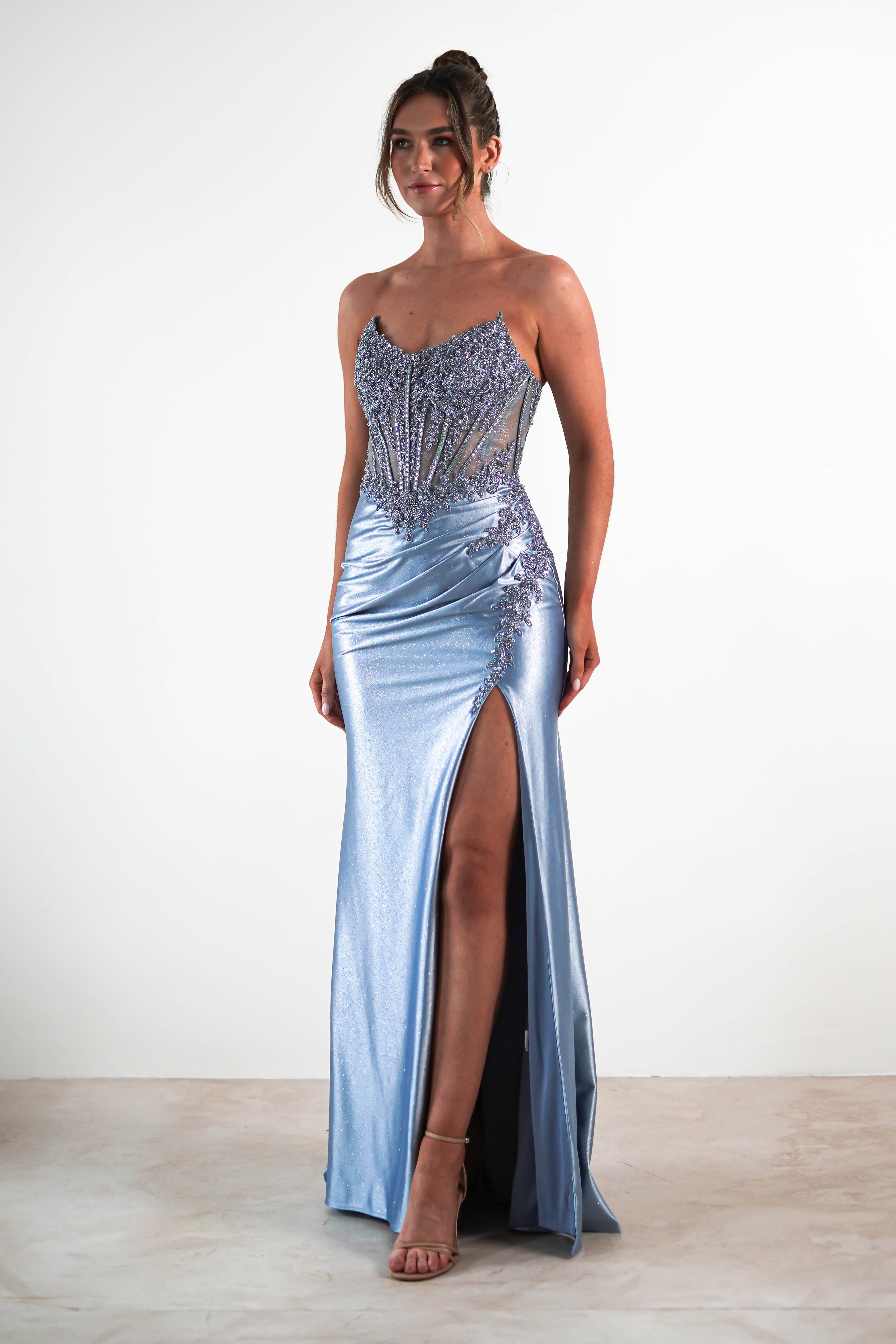 A woman wearing a shimmering, sequin embellished gown with a strapless bodice, thigh-high slit, and nude high heels stands against a plain white background. Intricate detailing highlights the elegant evening look.