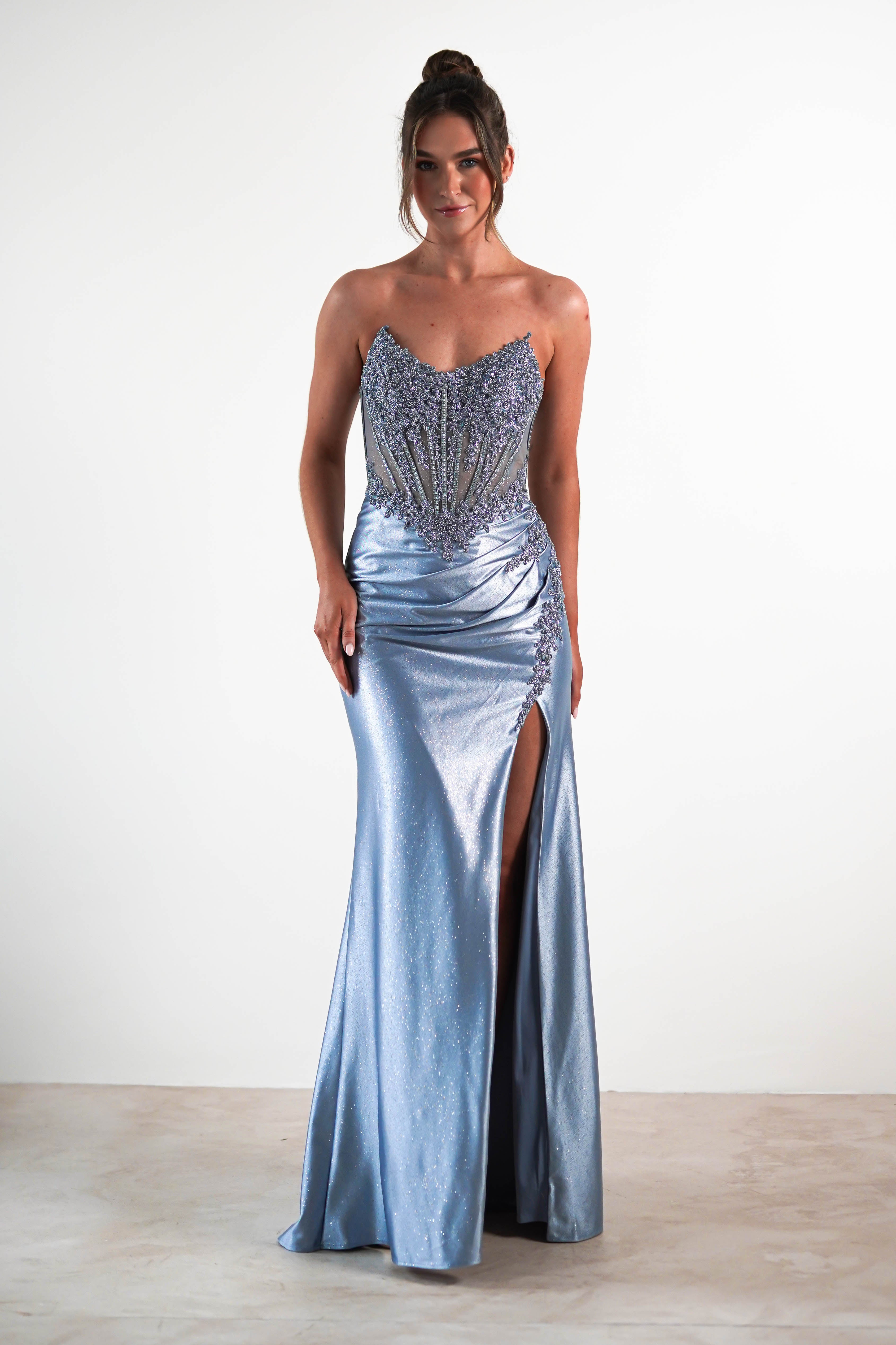 A woman in a shimmering sequin embellished gown with a beaded bodice, high slit, and corset midriff stands against a plain white background, smiling with her hair in an elegant updo.