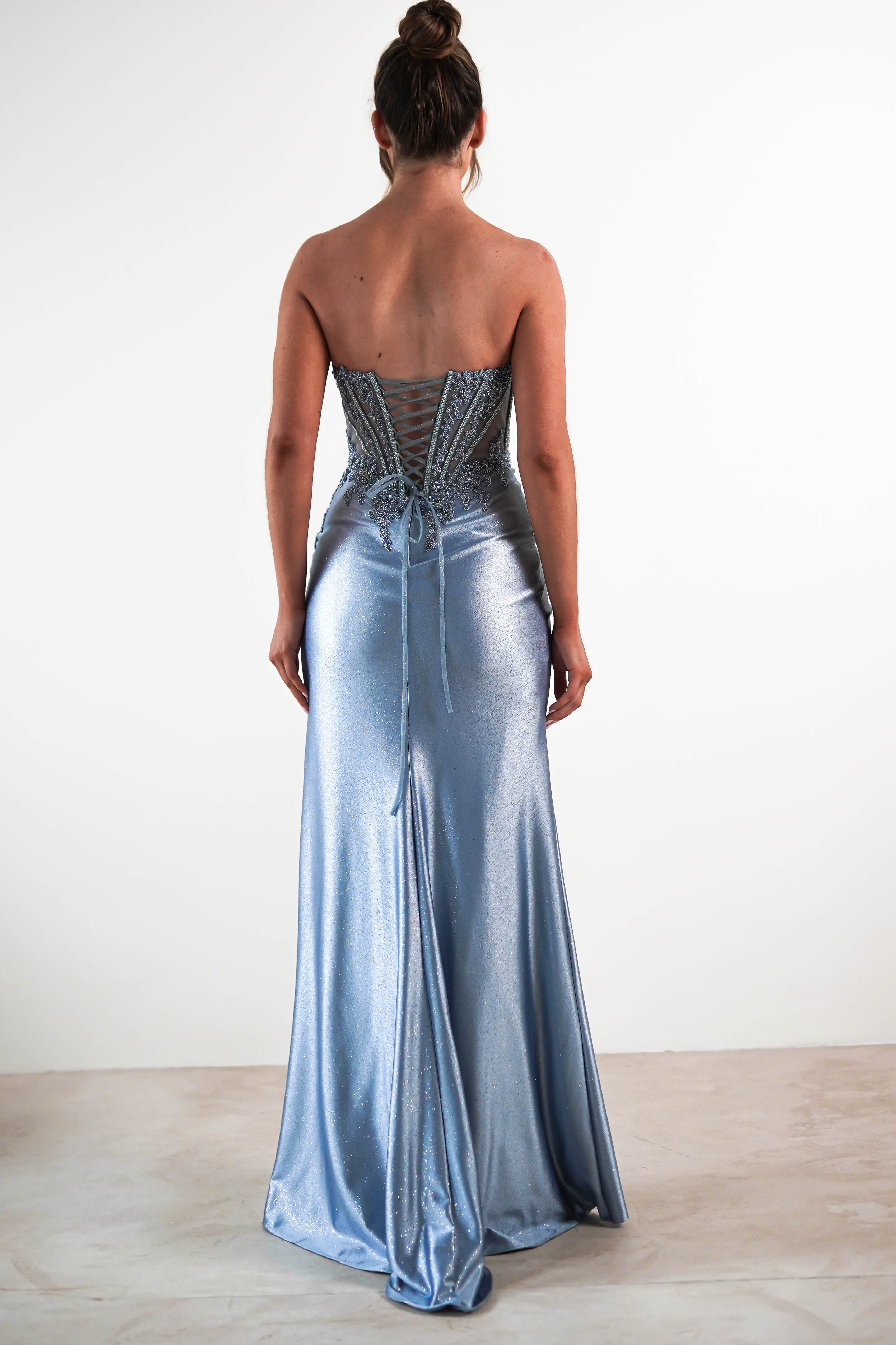 A woman stands facing away, wearing a light blue sequin embellished gown with lace-up detailing on the back and her hair styled in a bun. She is standing on a plain floor against a white background.