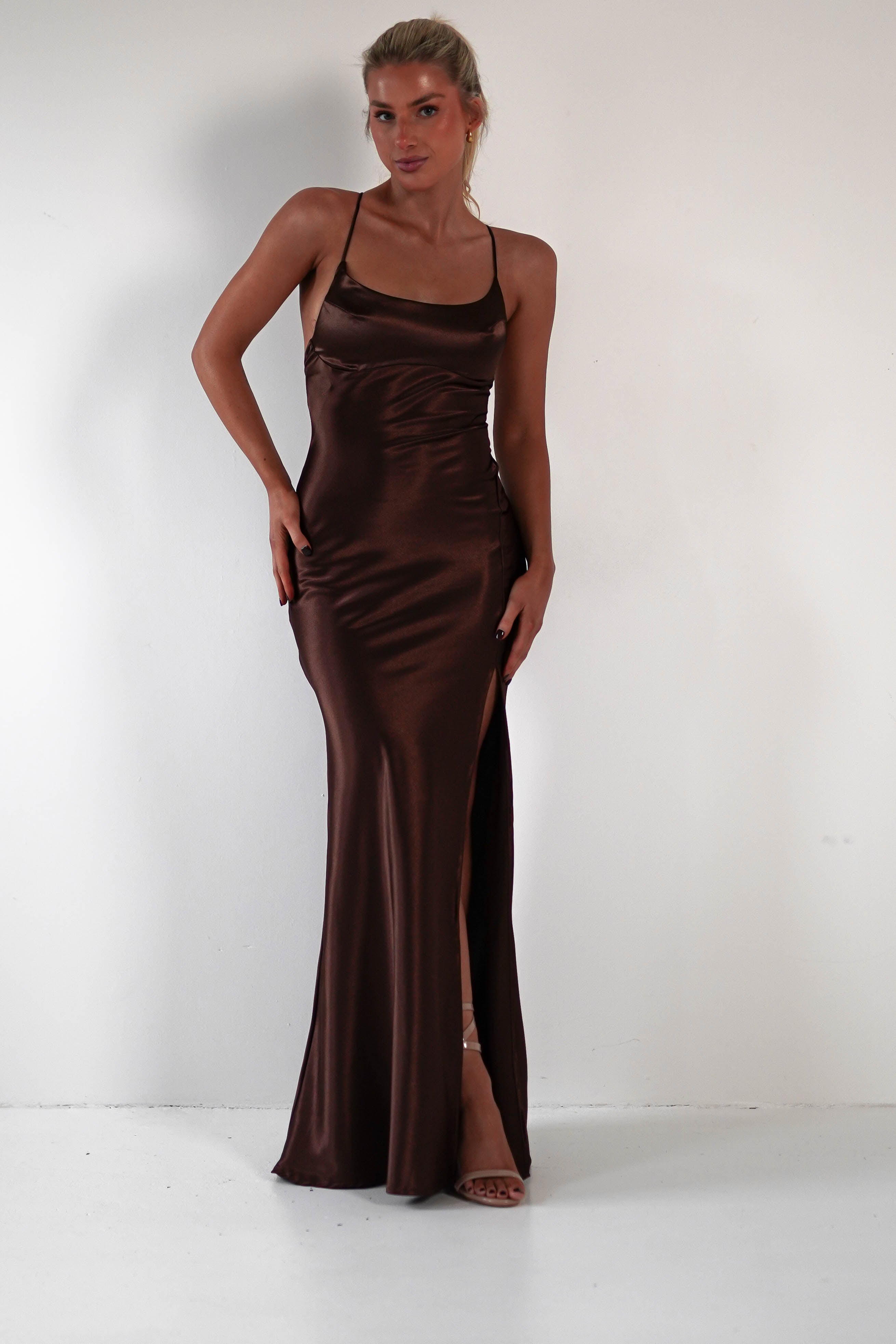 A person stands against a plain white background, wearing a long, brown satin gown with thin straps and a high slit that highlights its figure-hugging silhouette.