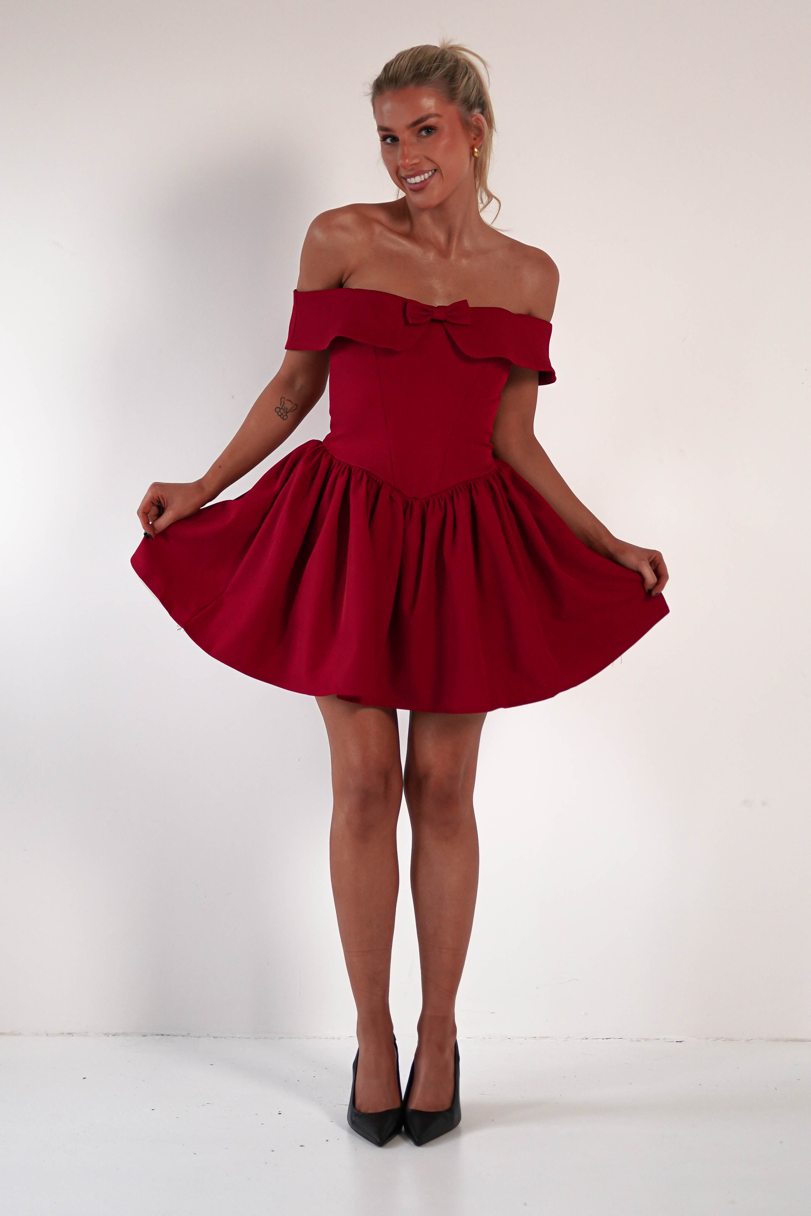 A person stands against a plain backdrop, wearing a red off-the-shoulder dress with a bow detail and flared, skater mini skirt. With a corset bodice accentuating their figure, they smile brightly while holding the dresss sides, paired elegantly with black heels.
