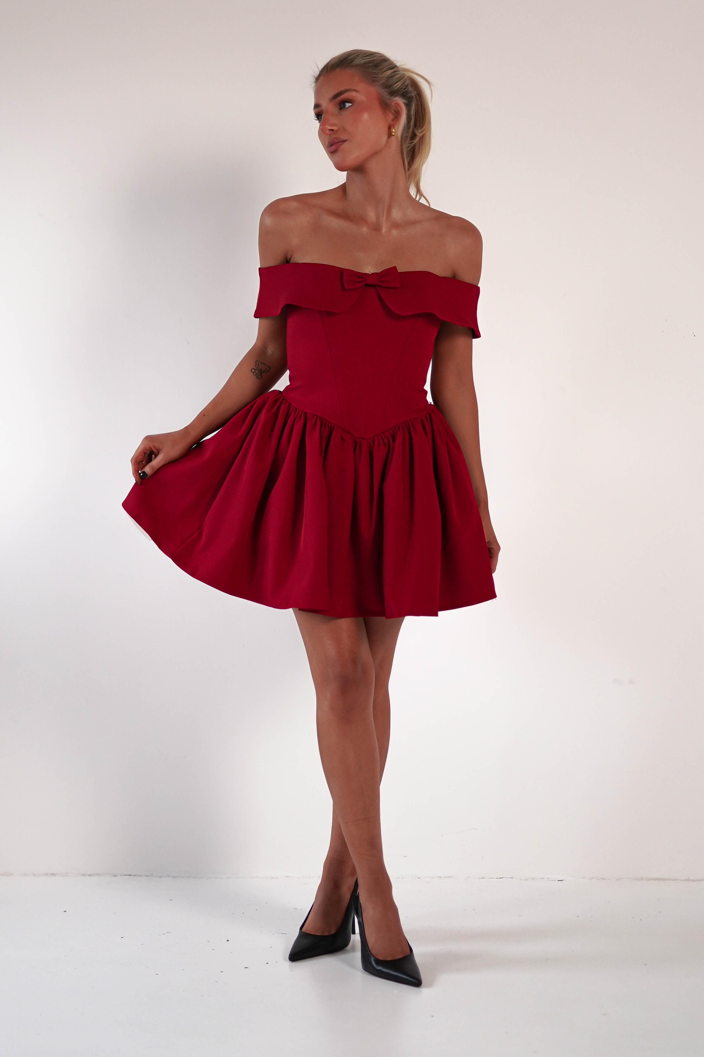 A person stands in a red, off-the-shoulder dress with a corset-shaped bodice and flared skirt, looking to the side. They are in a white room and wearing black heels, their right hand gently holding the dress to the side.