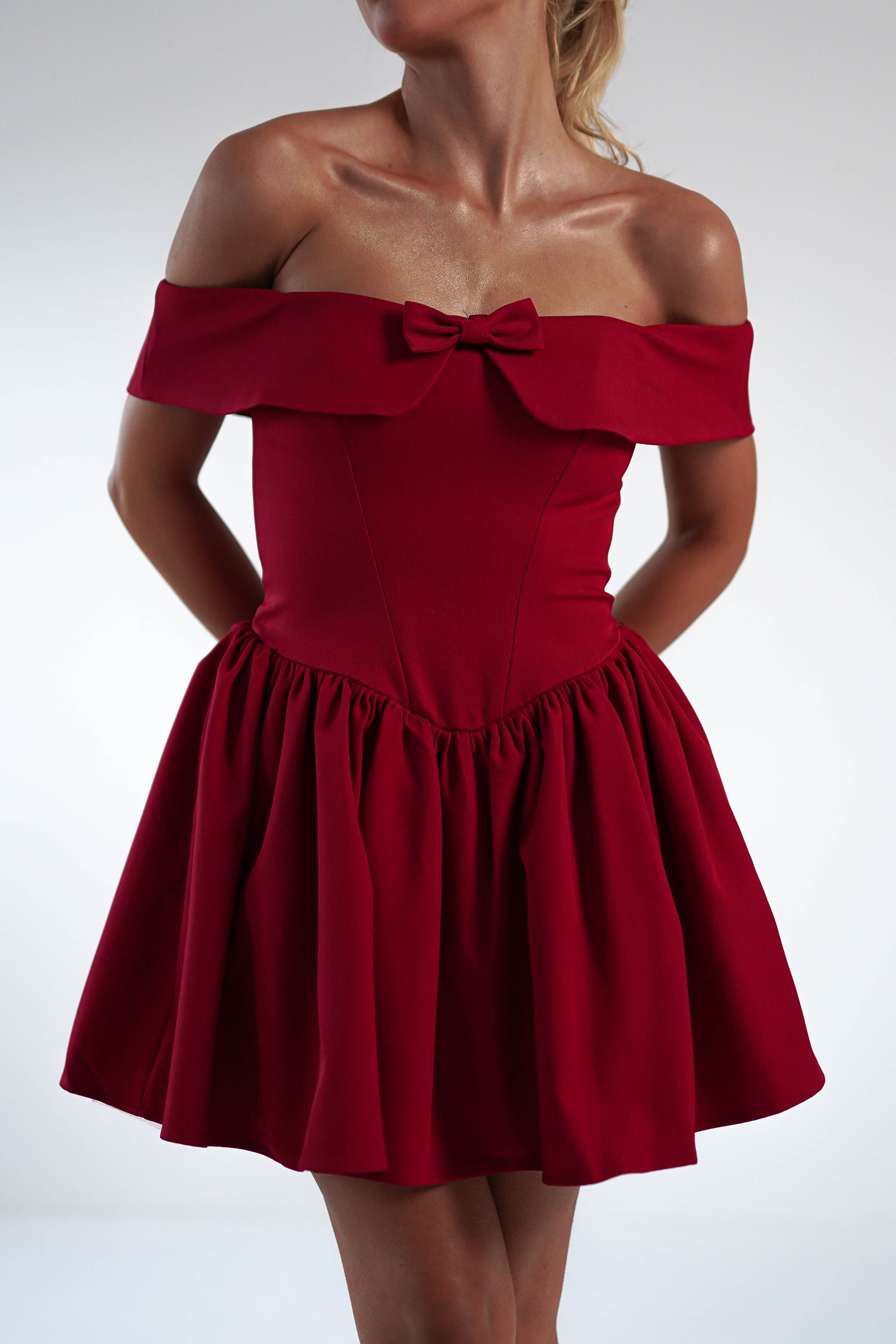 A person wearing a red off-the-shoulder dress with a bow on the neckline, posing against a plain background. The dress features a fitted corset bodice and flares into a playful skater mini skirt.