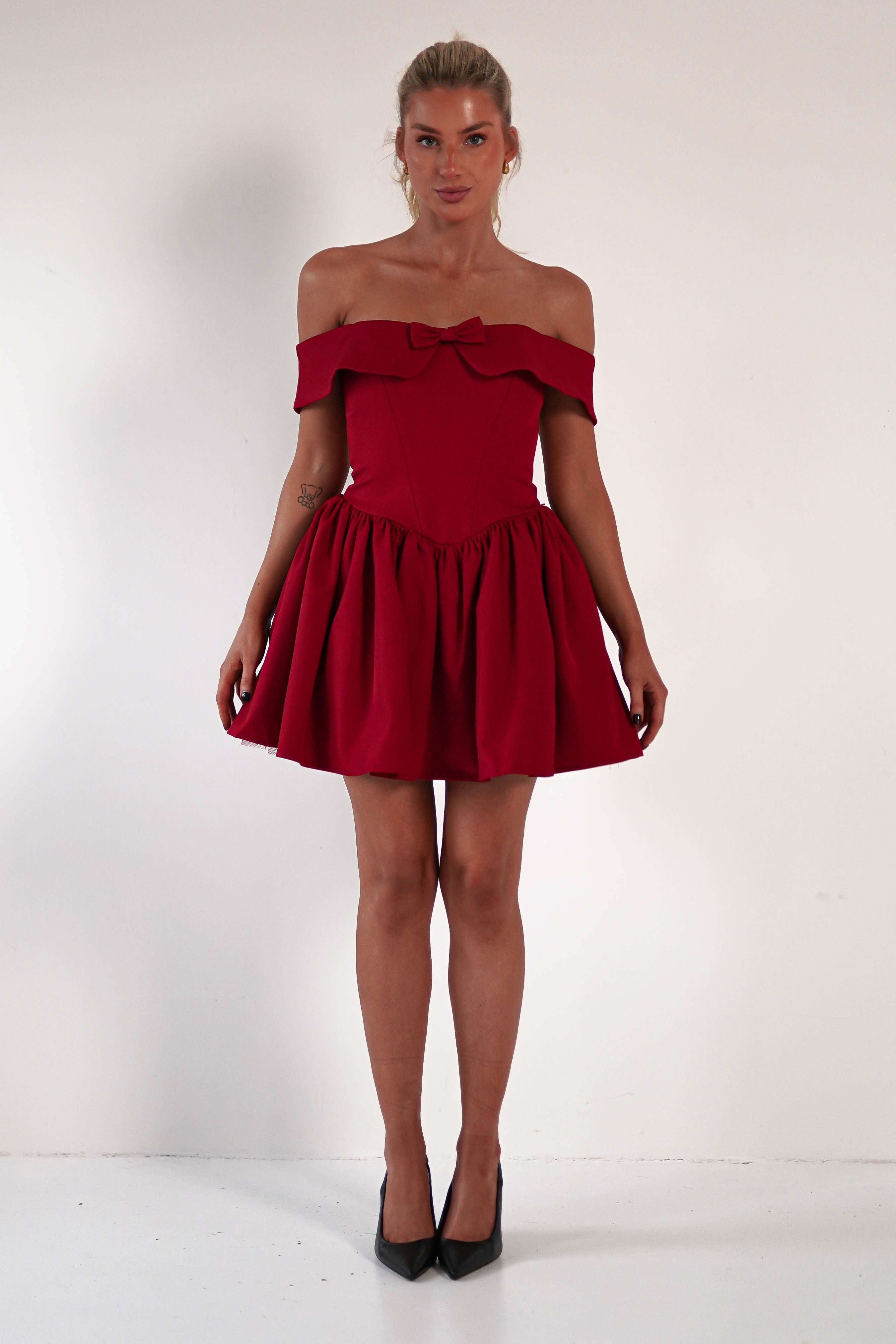 A person stands against a plain white background, adorned in an off-the-shoulder, bright red dress featuring a corset bodice and a bow detail. Their hair is tied back, and they complete the look with black high heels.