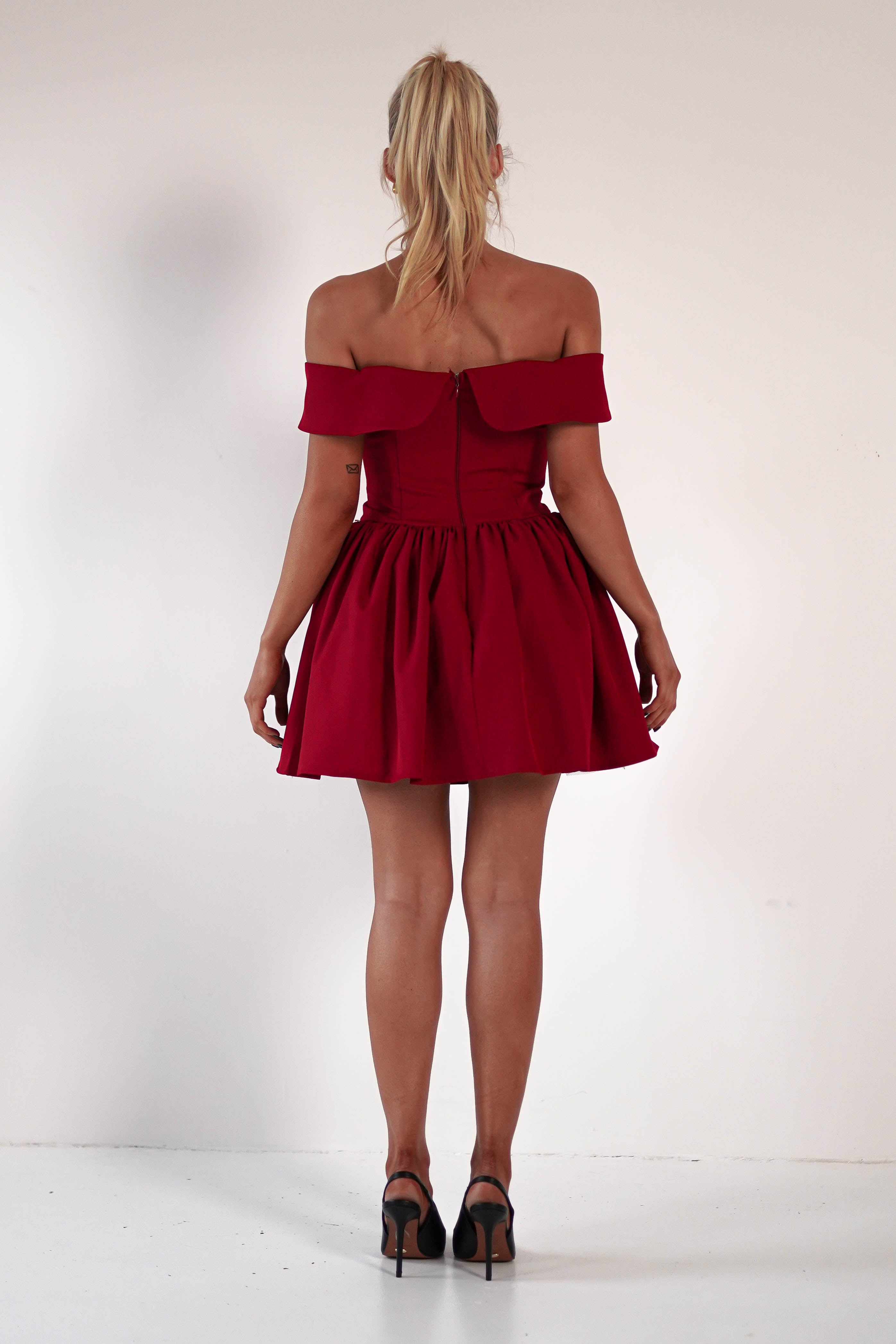 A woman with blonde hair in a ponytail stands with her back to the camera, wearing an off-the-shoulder red dress and black high heels. The skater mini dress features a fitted corset bodice and flares into a playful skirt against a plain white background.