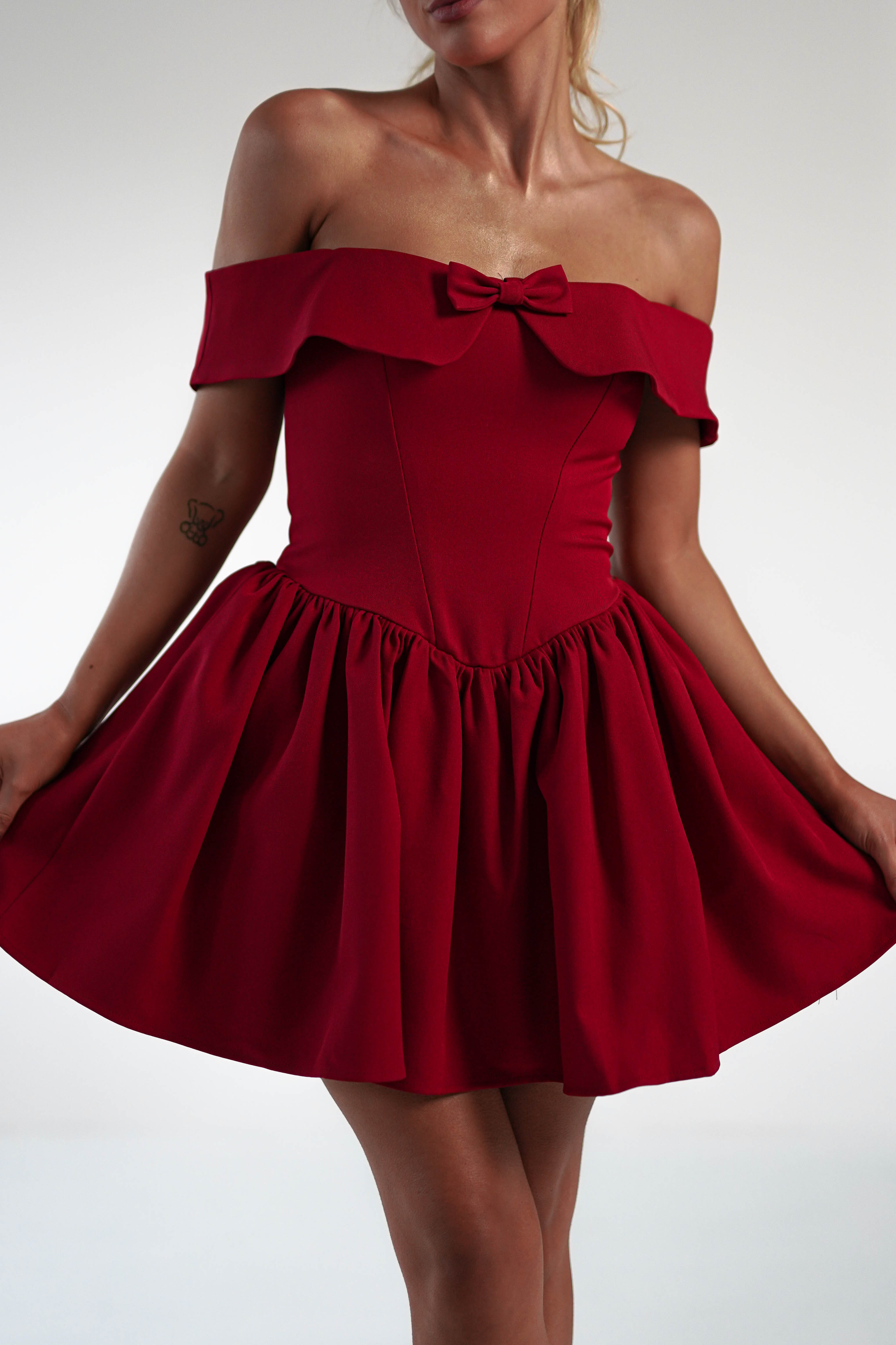 A woman dons an off-the-shoulder red dress with a short, skater mini skirt and a playful bow at the neckline. Her tattoo graces her left arm against a plain, light-colored backdrop.