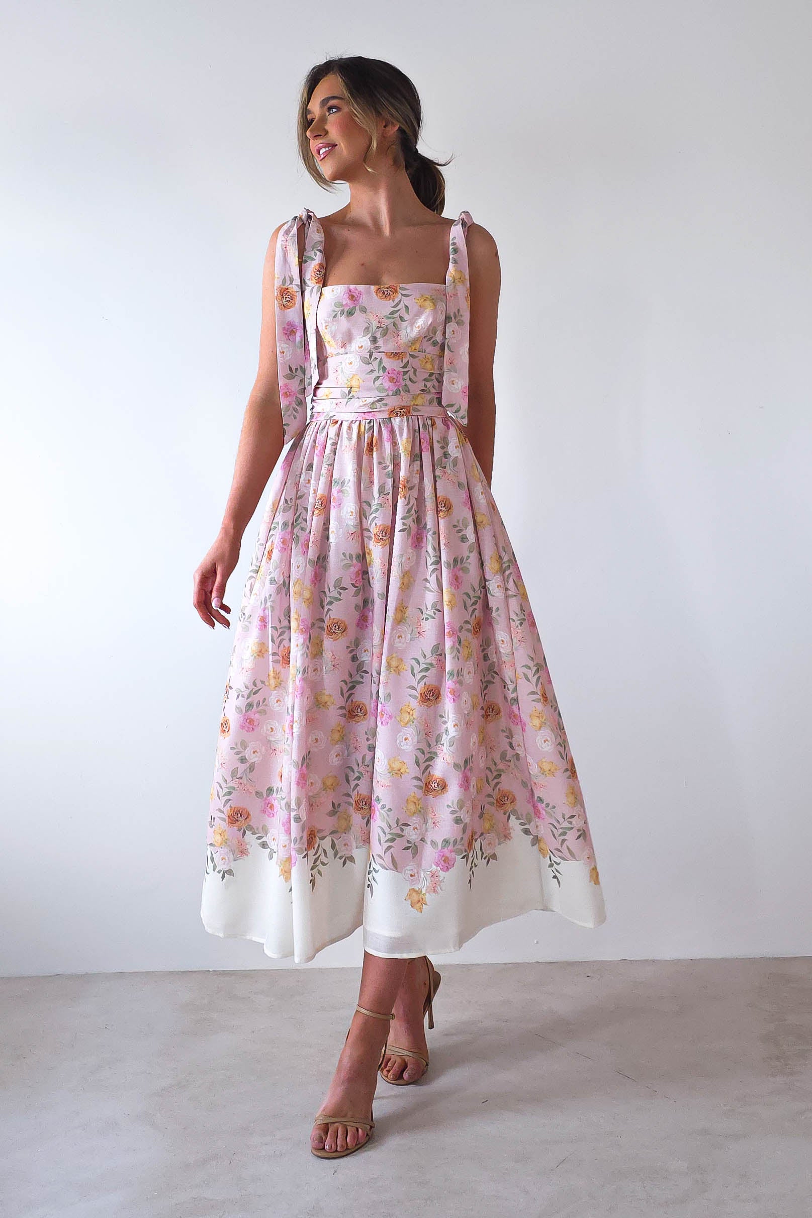 Woman wearing a floral pink and white summer print maxi dress with shoulder ties, looking to the side and smiling. The dress has a fitted bodice and flowing skirt. She stands against a plain white background, perfect attire for wedding guests, paired with beige high heels.