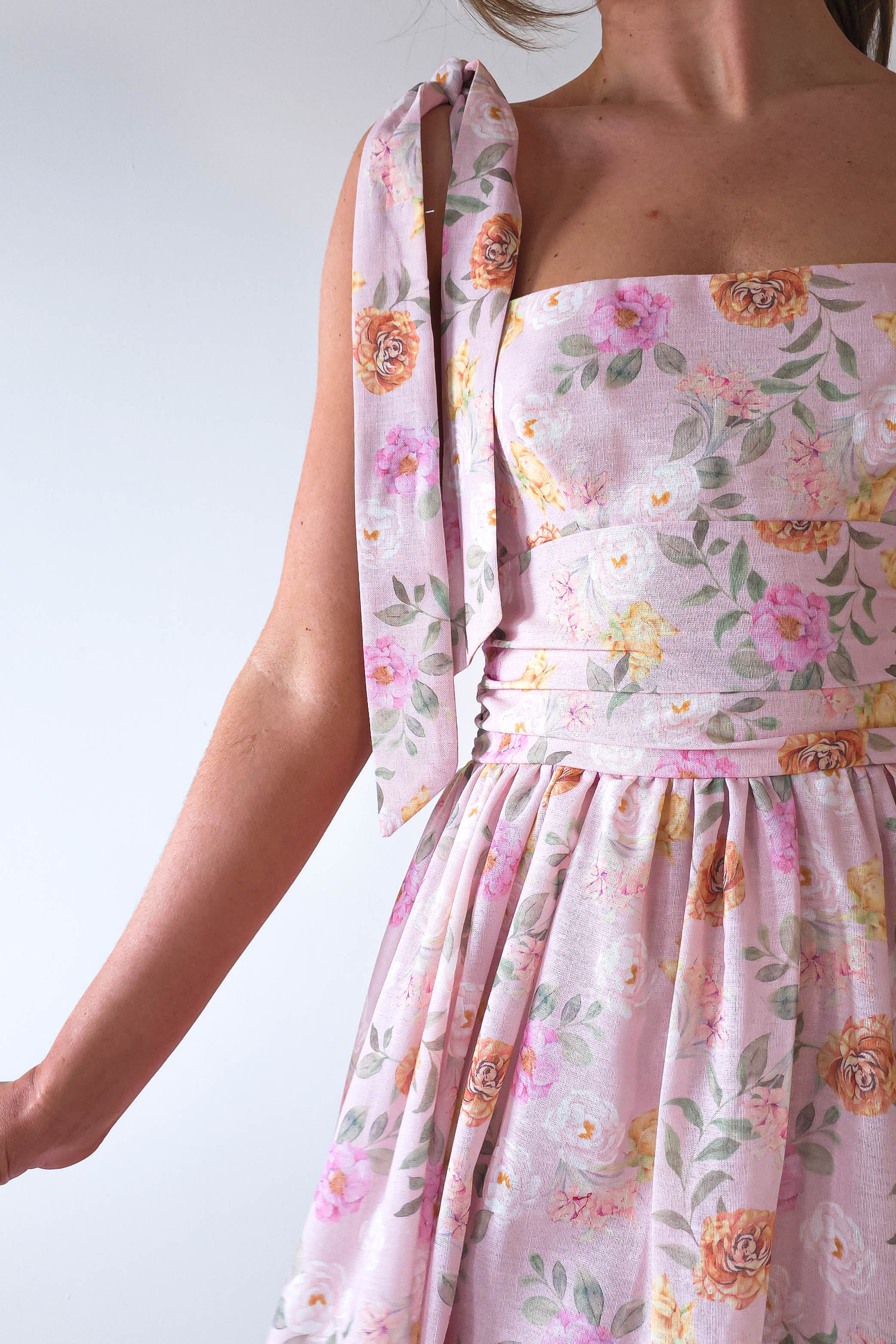 A person wearing a pink, floral-patterned summer print maxi dress with a shoulder bow stands against a plain white wall. The dress, perfect for wedding guests or other summer events, boasts flowers in shades of yellow and orange against its light pink background.