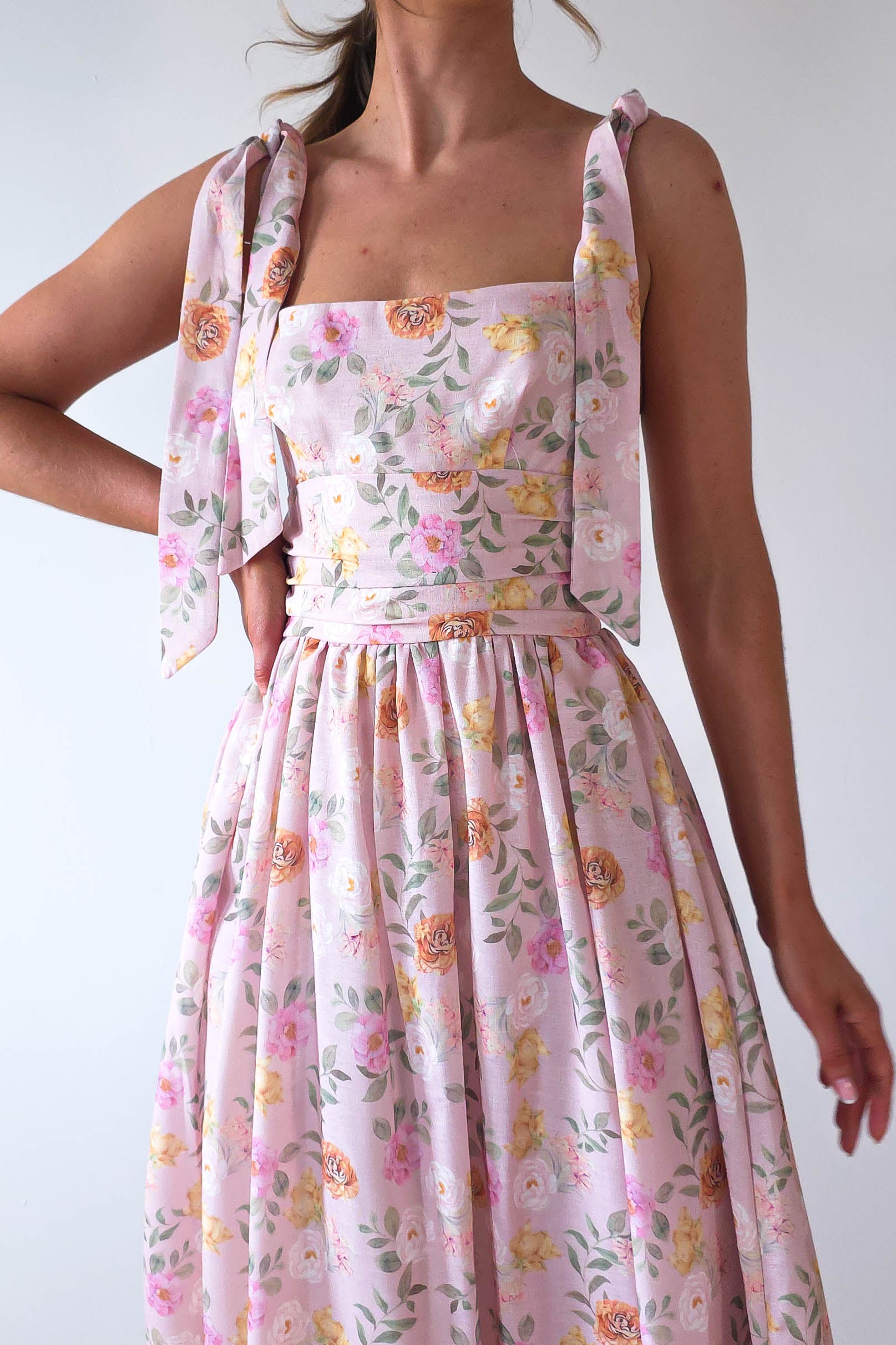 A woman wearing a pink floral dress with shoulder ties exudes effortless charm. With a square neckline and fitted bodice flowing into a maxi skirt, its perfect for summer events. The vibrant pattern of pink, yellow, and green makes it an ideal choice for wedding guests against any backdrop.