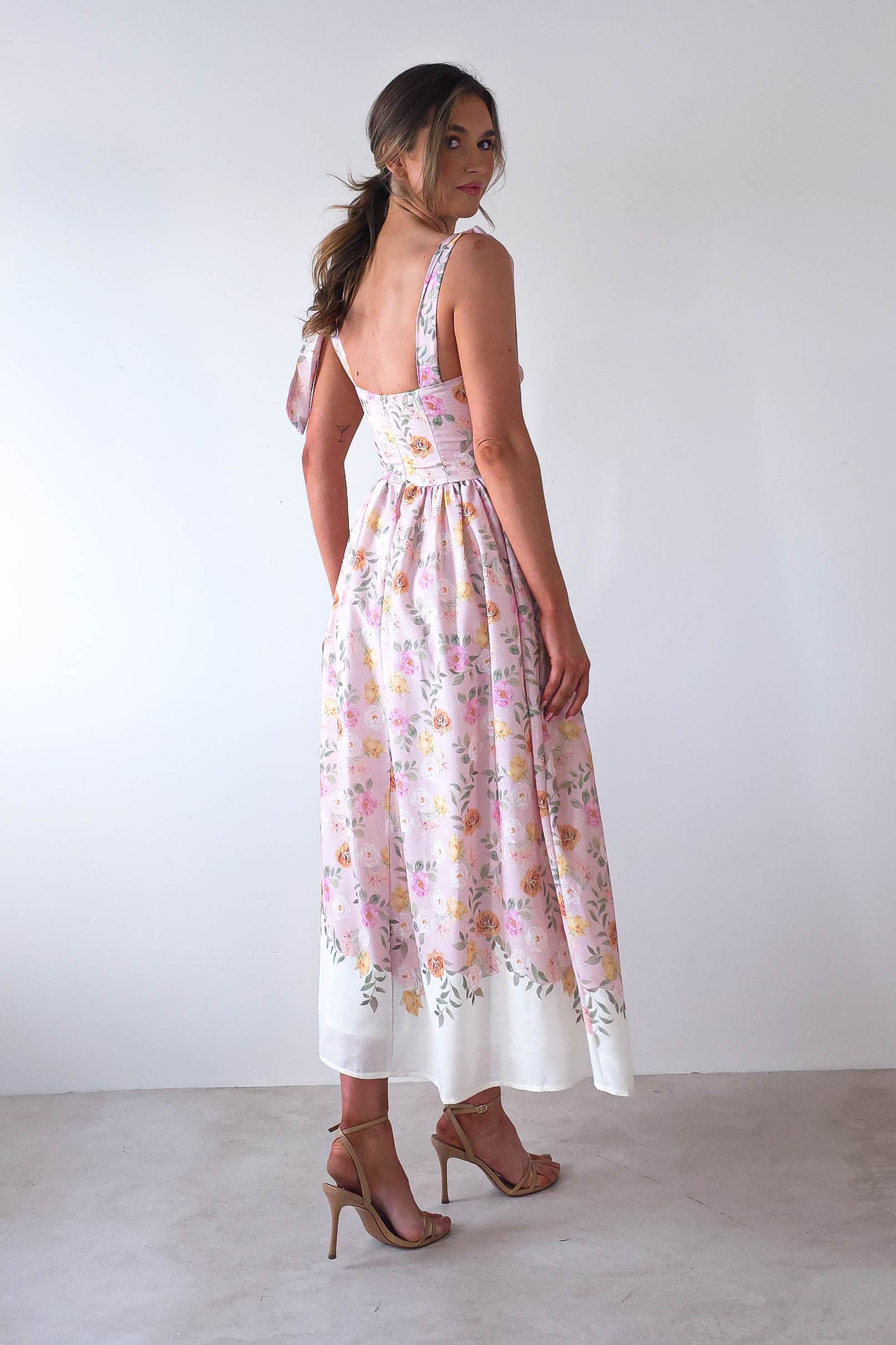 A woman stands facing sideways in a summer print maxi dress that flows to her ankles. Her hair is styled in a loose ponytail, and she wears high-heeled sandals, perfect for wedding guests or summer events. The background is plain white.