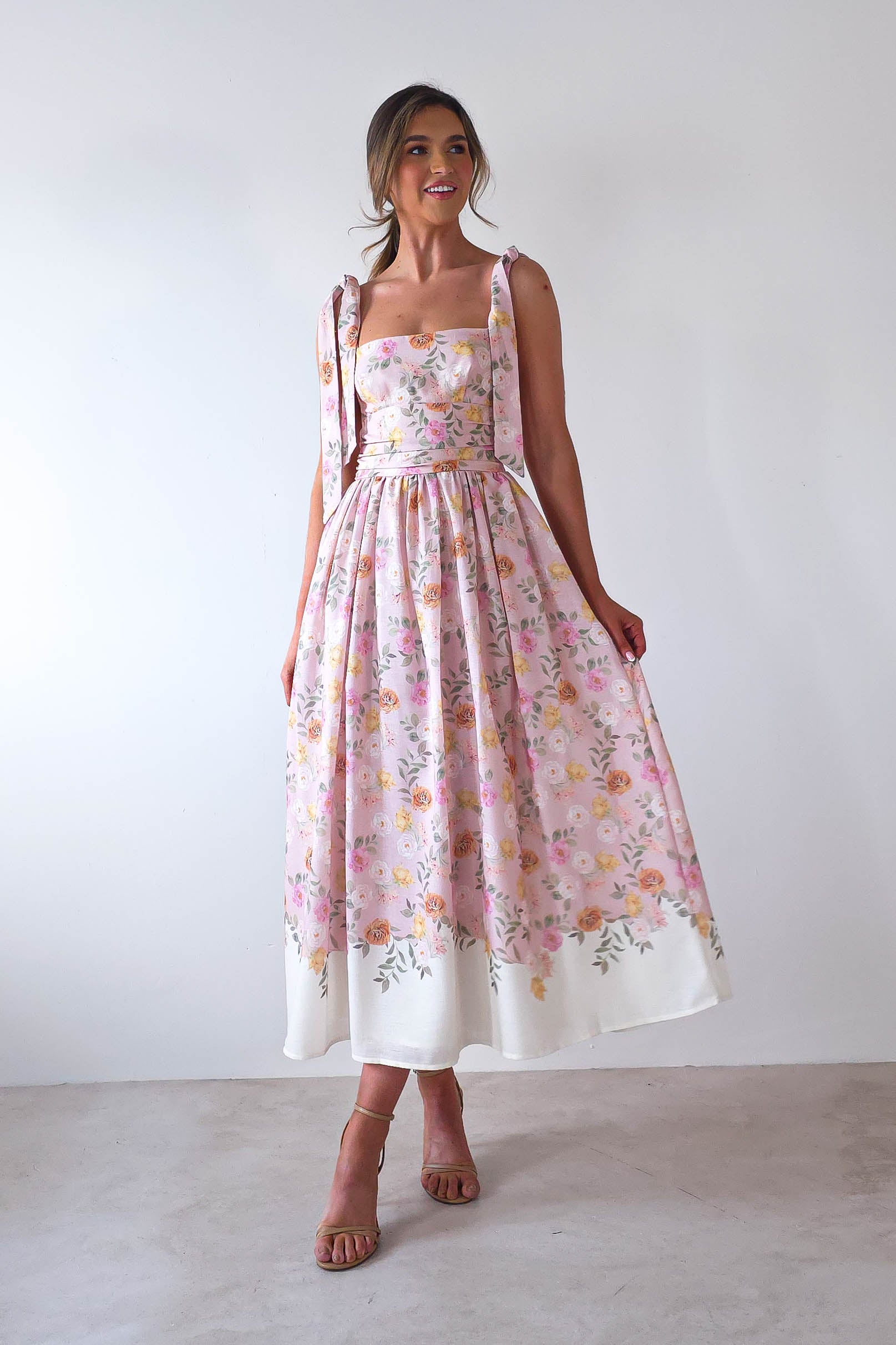A person wearing a sleeveless, summer print maxi dress with tie straps. The pink dress, adorned with yellow and white flowers, has a long, flowing skirt perfect for wedding guests. They stand against a plain white background, looking to the side with a smile.