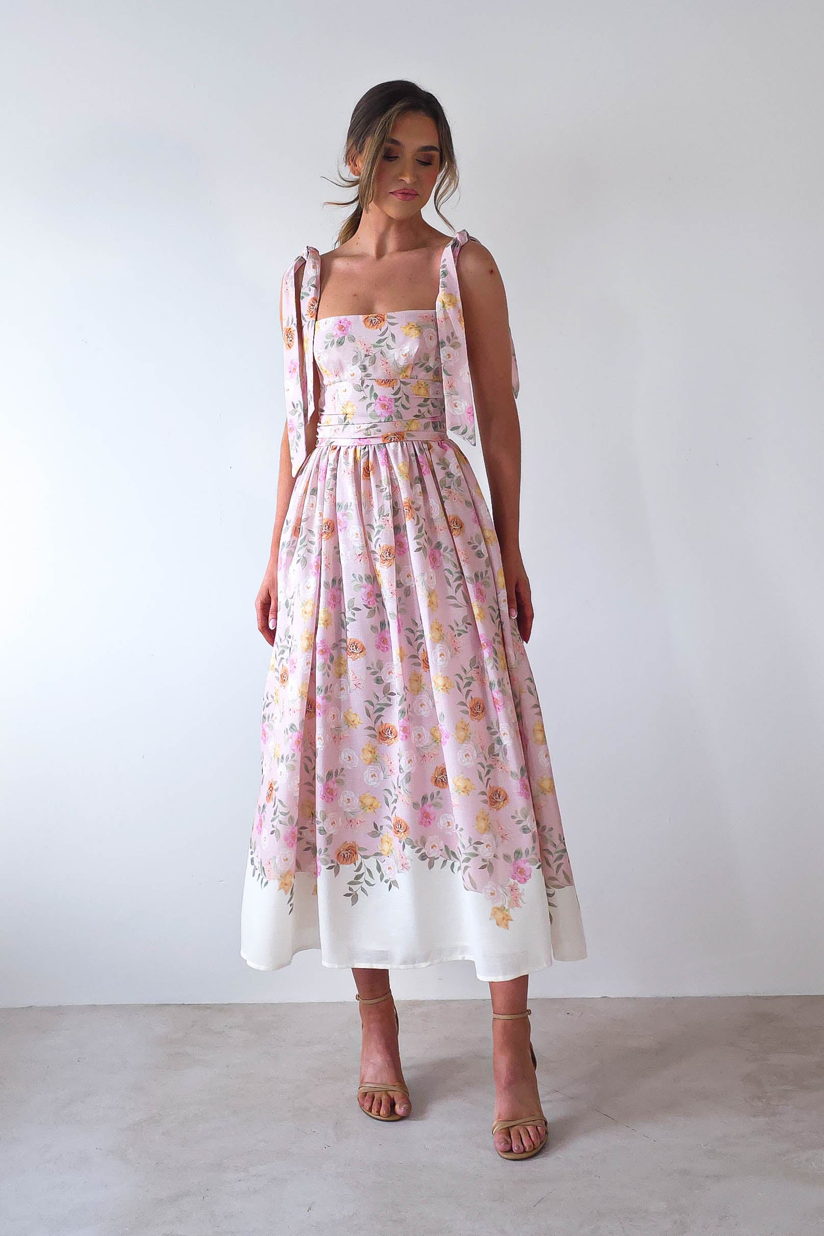 A woman in a floral pink and white dress with a fitted bodice and full skirt stands against a plain white background, embodying the elegance of summer events. Her high heels complement her look as she gazes downward, hair styled in a loose updo perfect for wedding guests.