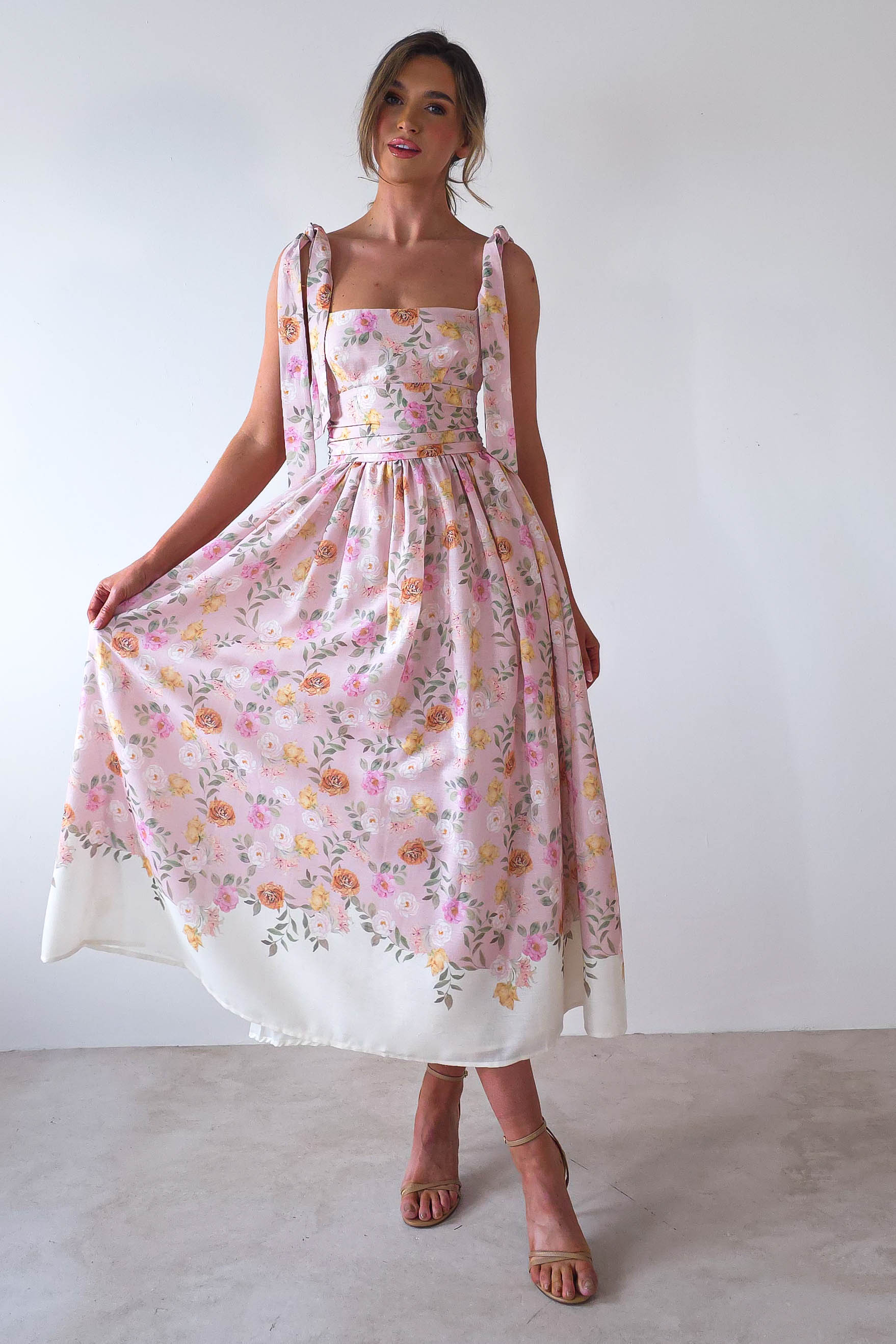 A woman in a floral pastel pink dress with a square neckline and tie straps poses elegantly in a light room. She holds the skirt, highlighting its summery pattern—an ideal choice for summer events. She wears strappy nude heels, and her hair is styled in a loose updo, perfect for wedding guests.