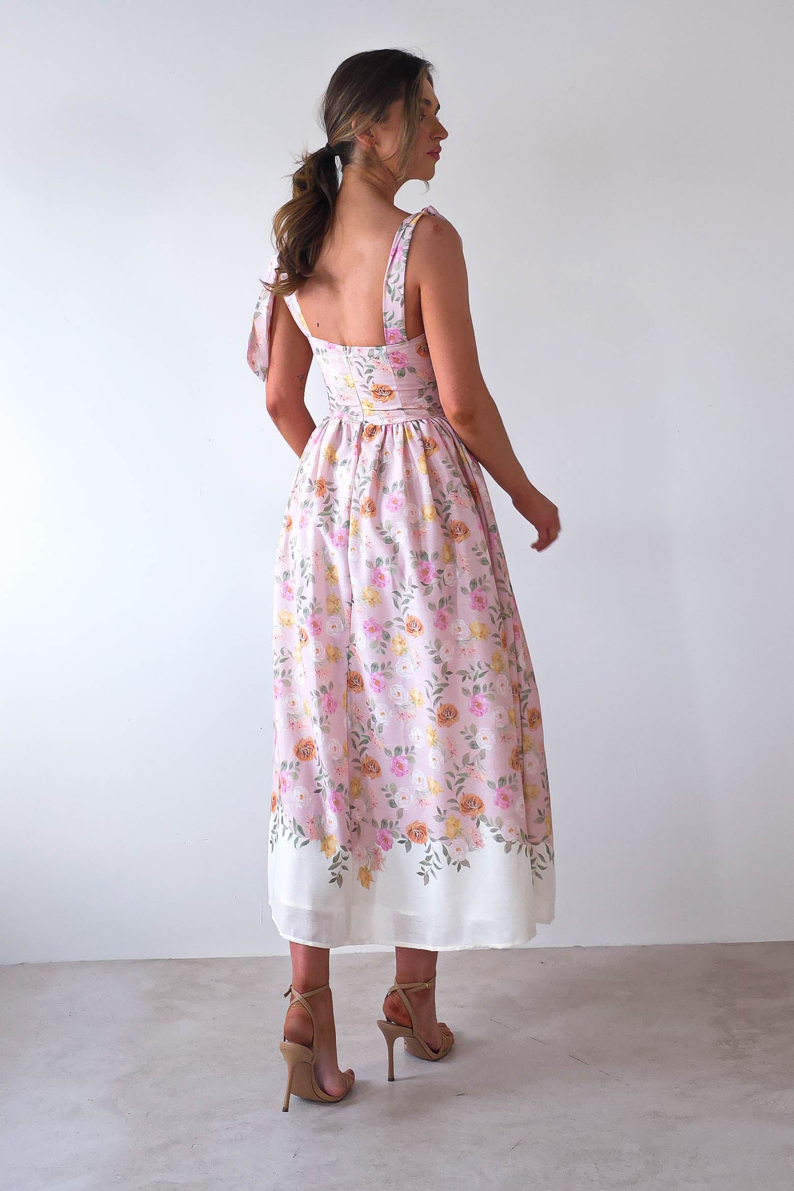 A woman in a pink floral dress stands facing a white wall, perfect for summer events. The dress, resembling a summer print maxi style, has a fitted bodice and flows to mid-calf with a white floral border. She wears high heels and her hair is tied back in a loose ponytail.