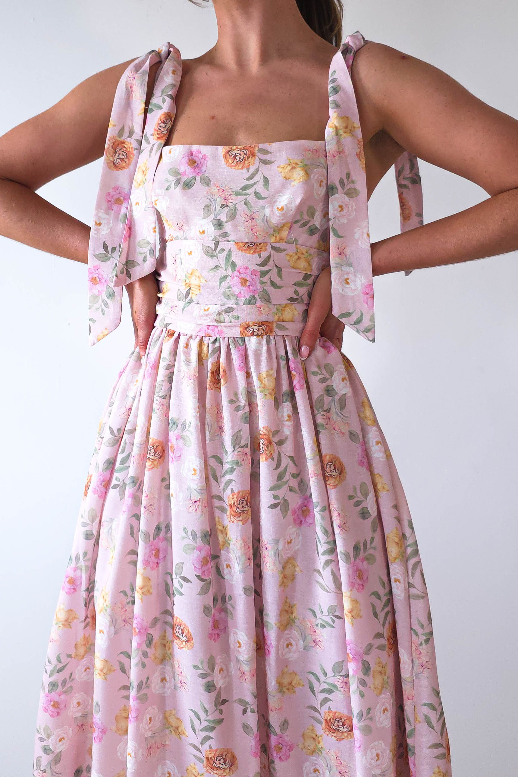 A person wearing a pink floral dress with tied shoulder straps stands with hands on hips. The summer print maxi dress features a square neckline and a flowing skirt adorned with roses and leaves, perfect for wedding guests. The background is plain white.