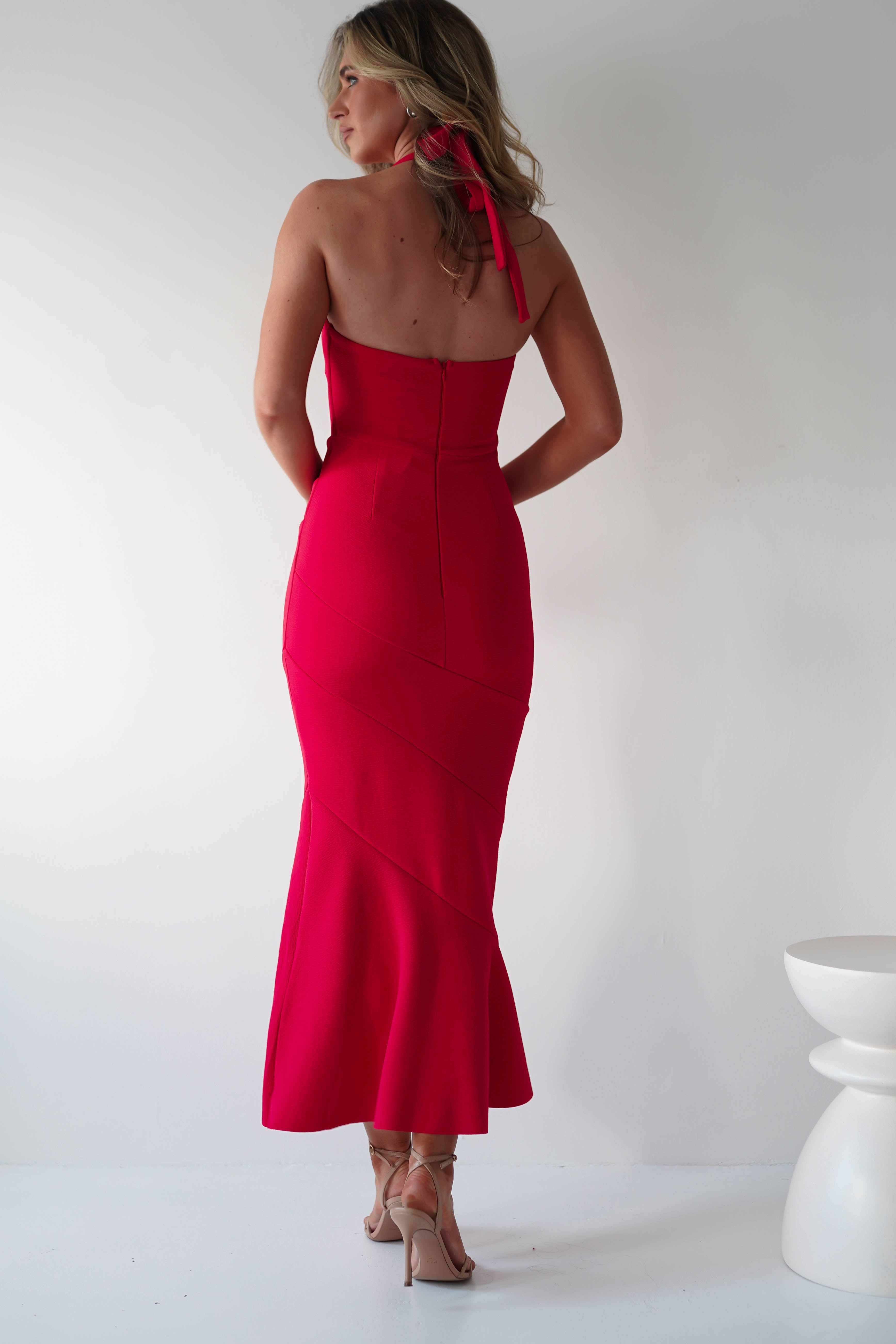A woman in a halterneck red midi dress and nude high heels stands with her back to the camera. Her shoulder-length blonde hair cascades elegantly, creating a perfect silhouette against the white backdrop, exuding charm fit for an unforgettable wedding guest.