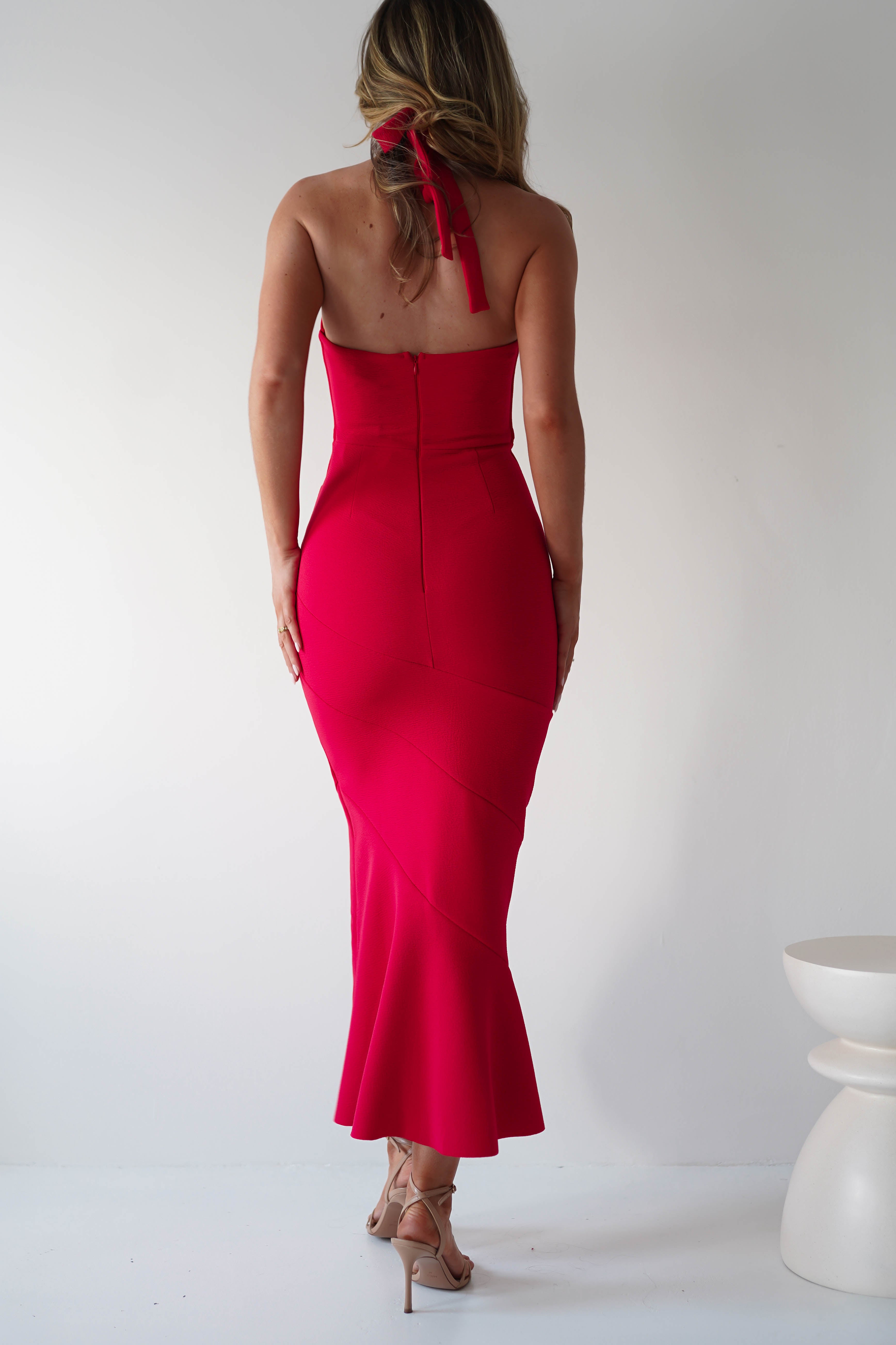 Rear view of a person with long hair in a striking red halterneck midi dress and beige high heels, standing against a plain white background. They are looking slightly to the side with one hand relaxed by their side, poised perfectly as an elegant wedding guest.
