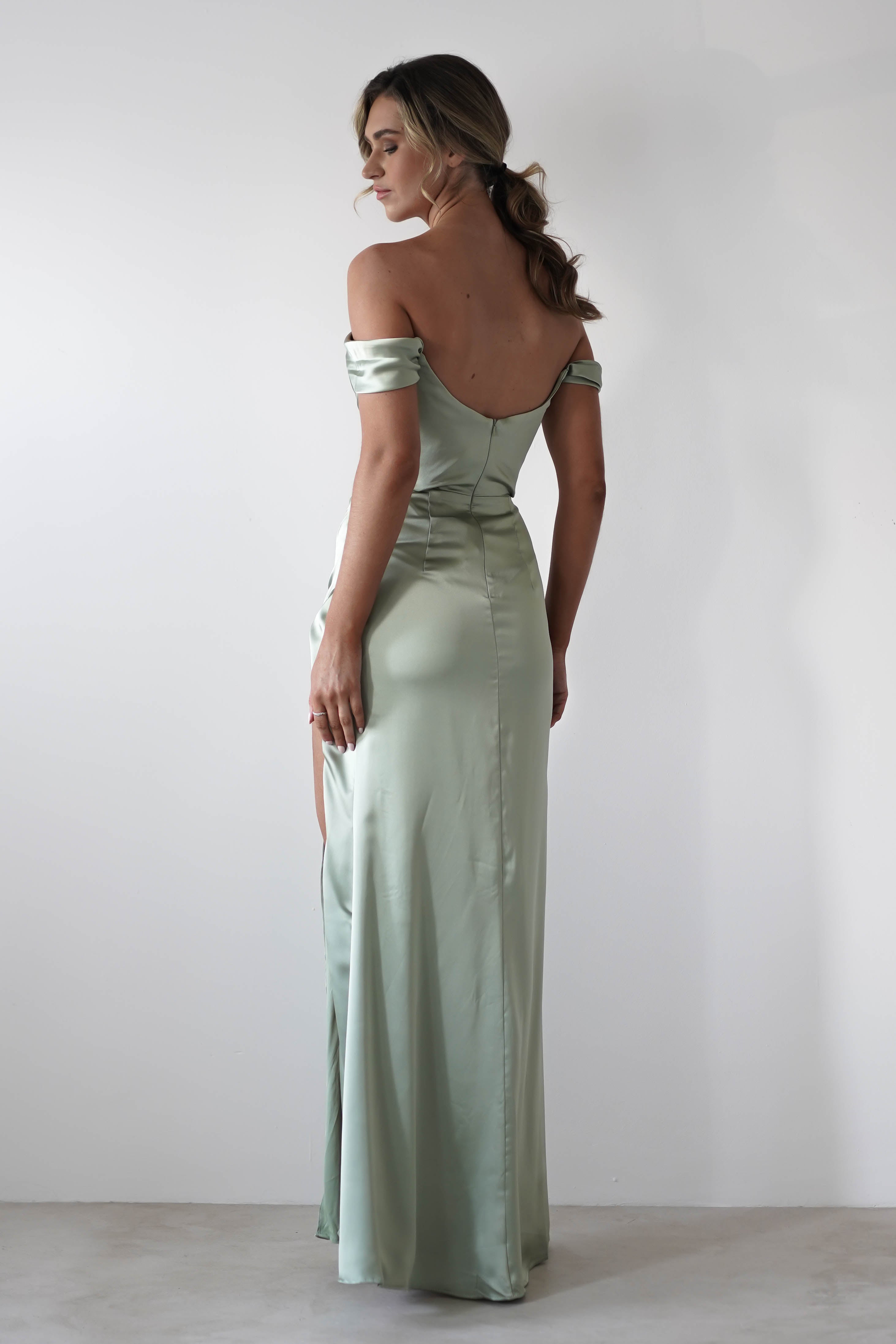 A woman stands facing away, wearing a long, light green thick satin fabric dress with a fitted waist and low back. The off-shoulder gown features a subtle cowl neckline, and her loose waves fall softly against the plain white background.