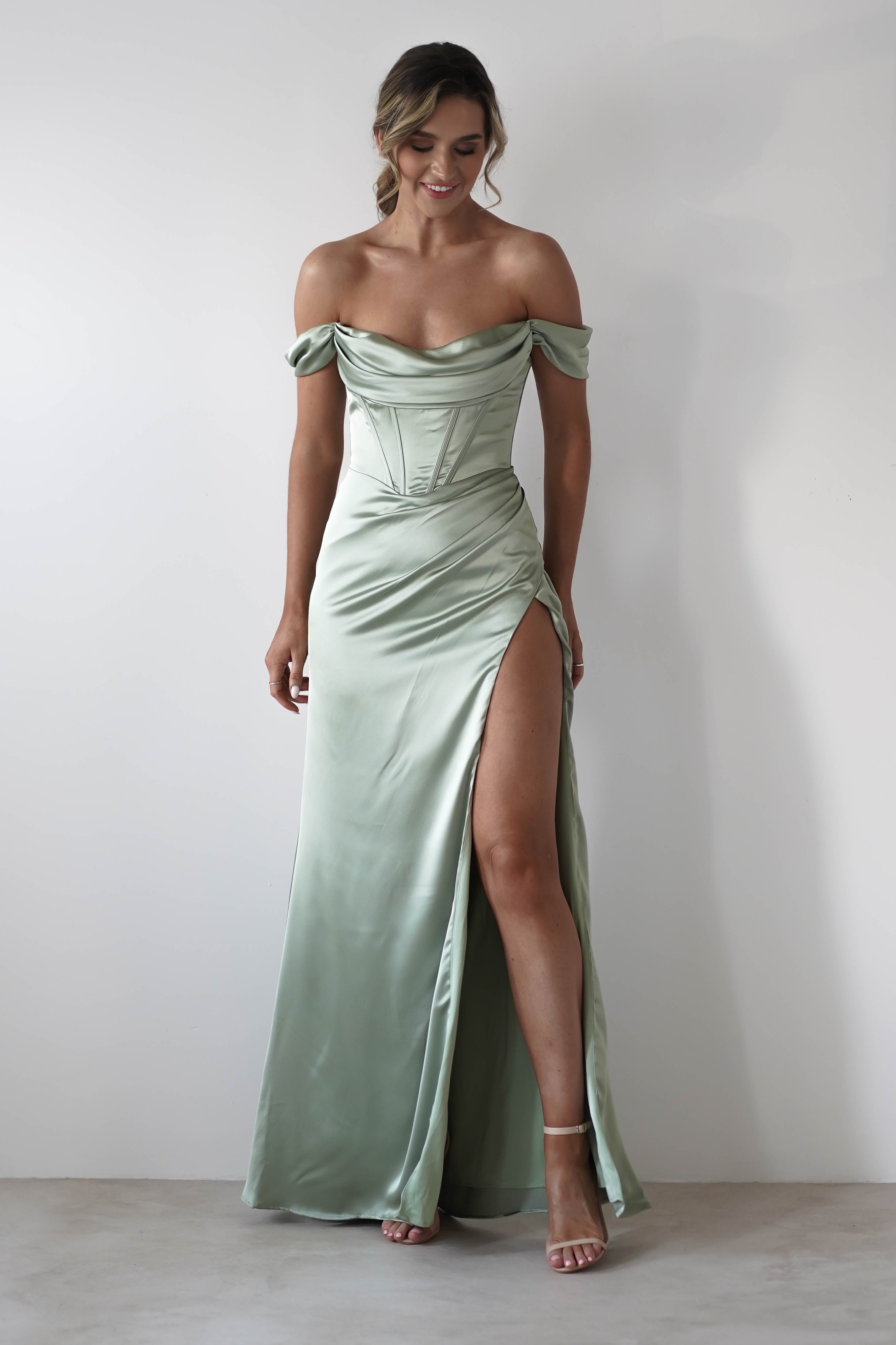 A woman in a light sage green thick satin fabric off-the-shoulder gown with a corset bodice and high thigh slit, featuring a chic cowl neckline, stands against a white background wearing strappy high heels.