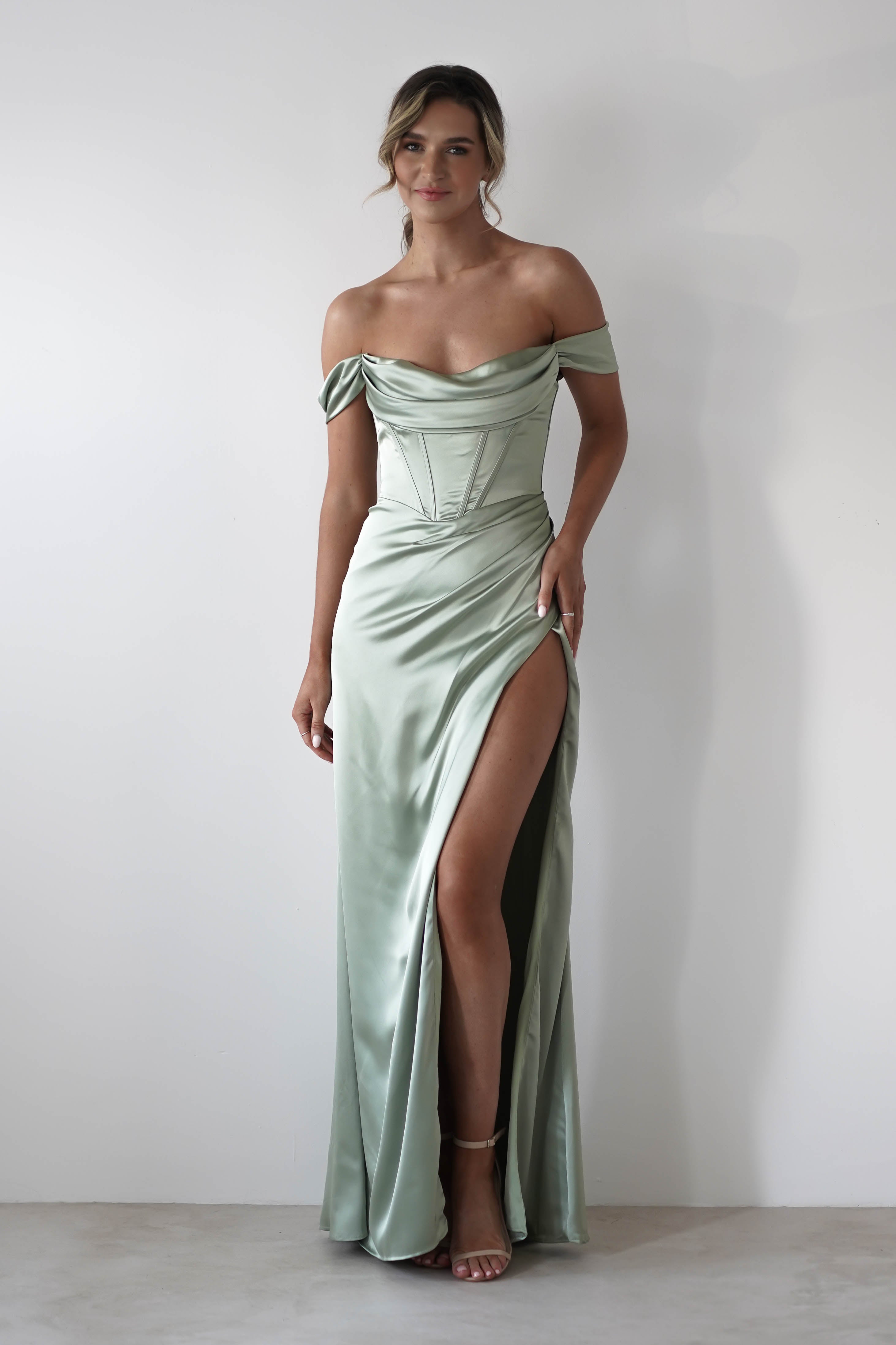 Woman wearing a light green, off-shoulder satin gown with a thigh-high slit and corset style bodice, standing against a plain white background.
