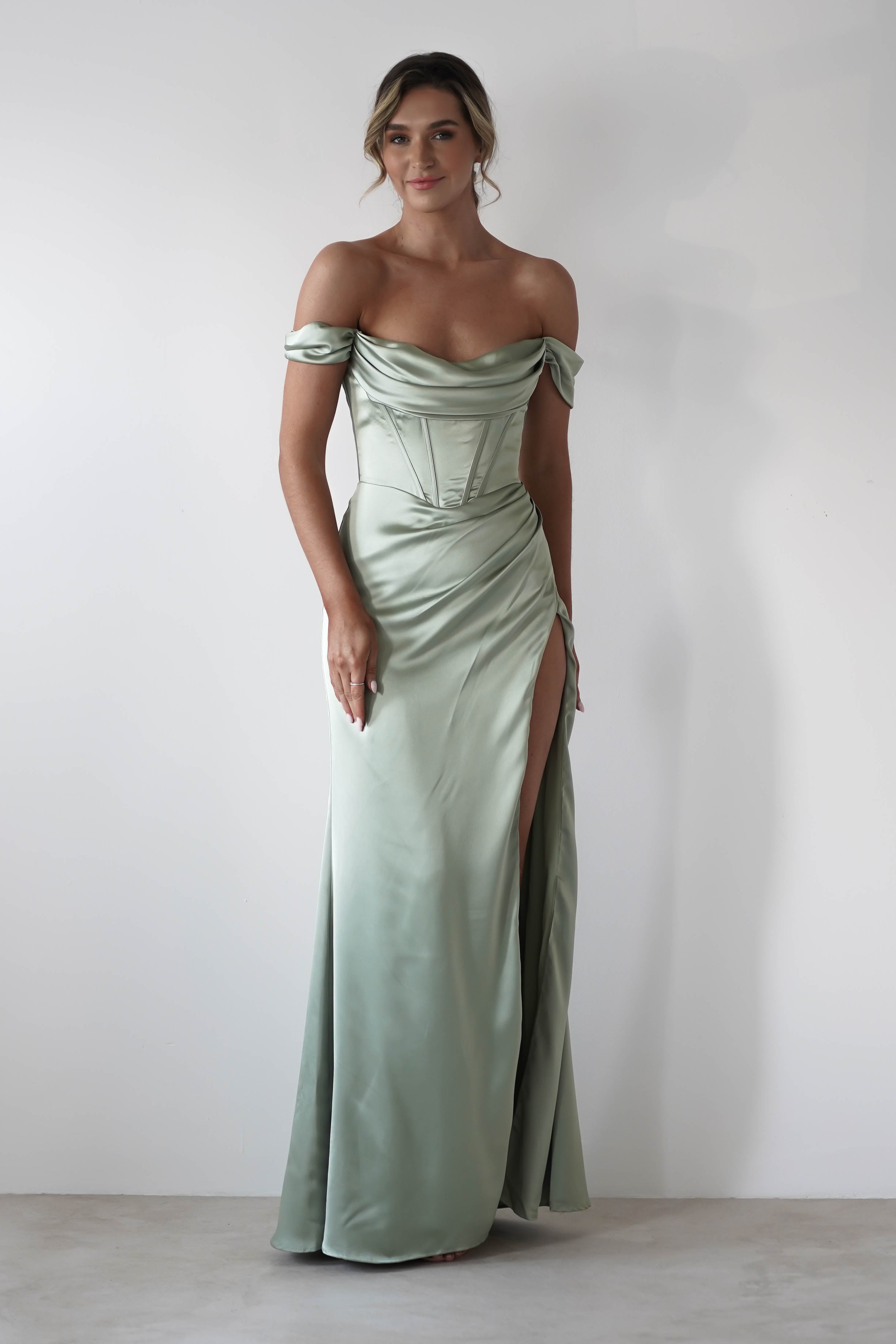 A woman in an off-the-shoulder, light green satin gown with a cowl neckline and thigh-high slit stands against a plain white background, smiling softly with her hair styled in an updo.