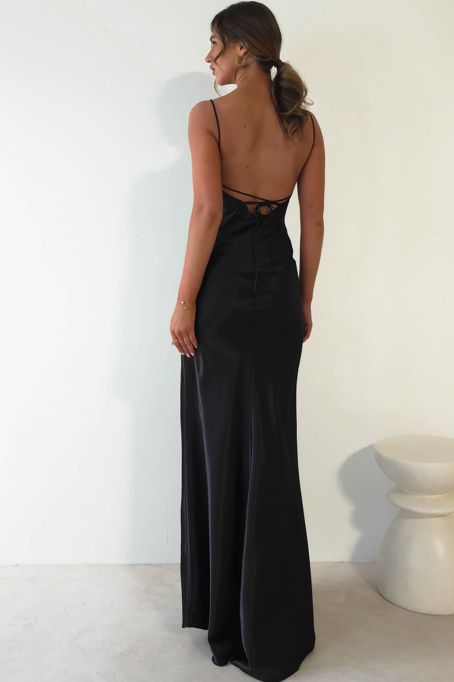 A woman stands with her back to the camera, wearing a black, satin gown with a figure-hugging silhouette and thin straps. Her hair is styled in a loose ponytail. She stands near a light-colored, rounded stool against a plain wall.