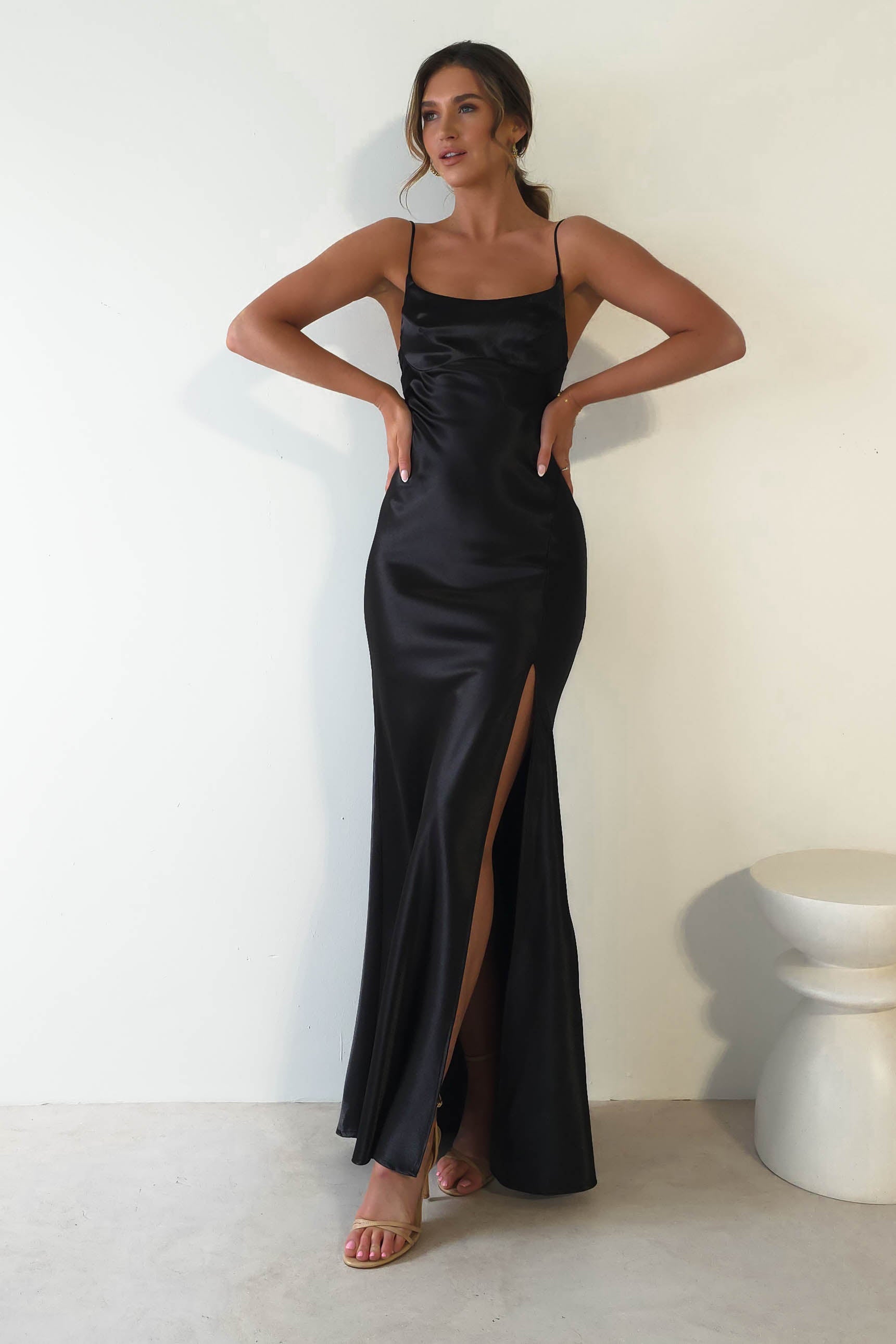 Woman in a sleek black satin evening gown with a figure-hugging silhouette, featuring thin straps and a high slit, standing with hands on hips in a minimalistic room. The dress elegantly drapes to the floor, paired with strappy heels. A white stool is visible in the background.