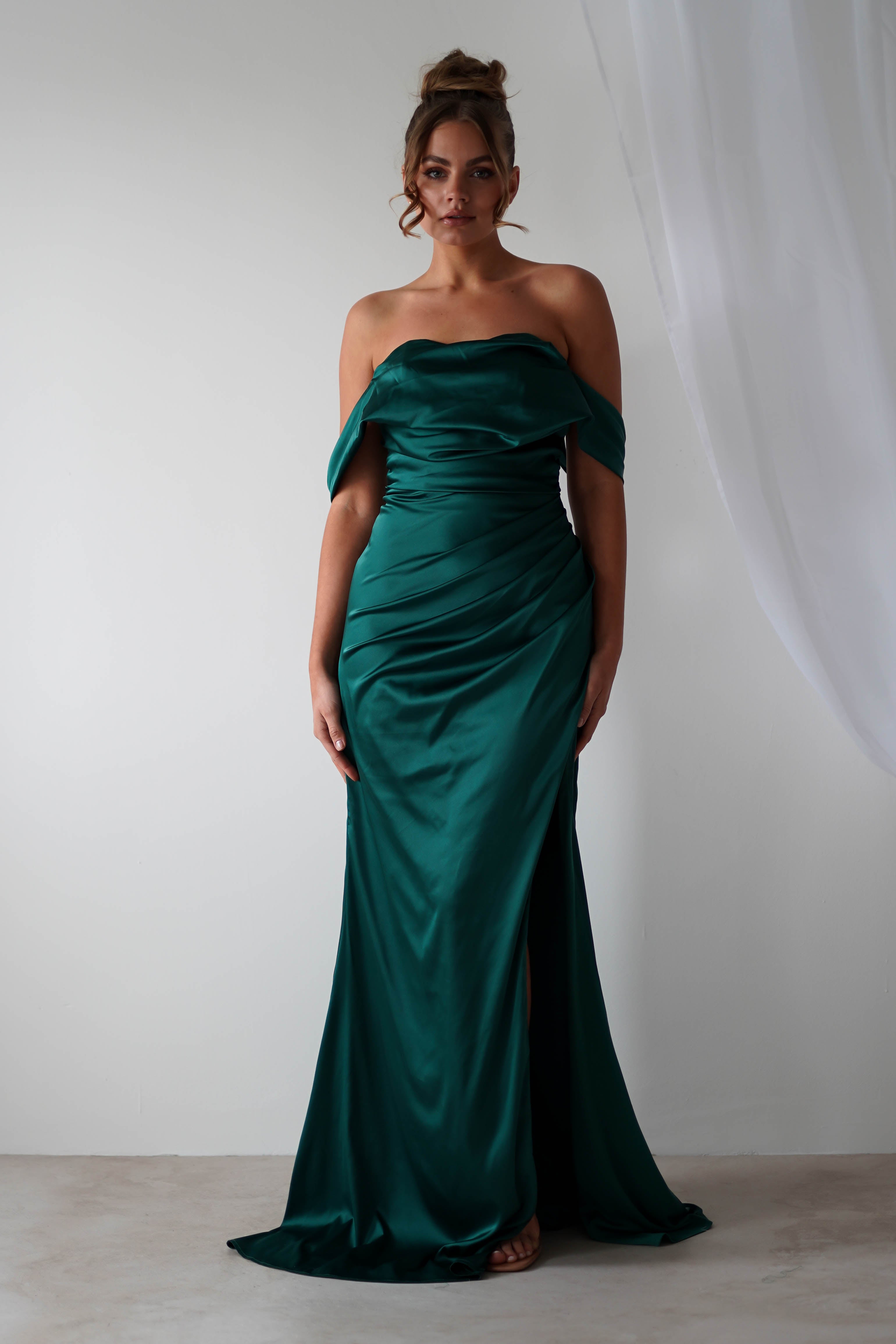 A woman wearing an off-the-shoulder gown made of rich satin material stands against a plain white background with sheer white drapery to the side. Her hair is styled in an elegant updo, perfect for bridesmaids or special occasions.