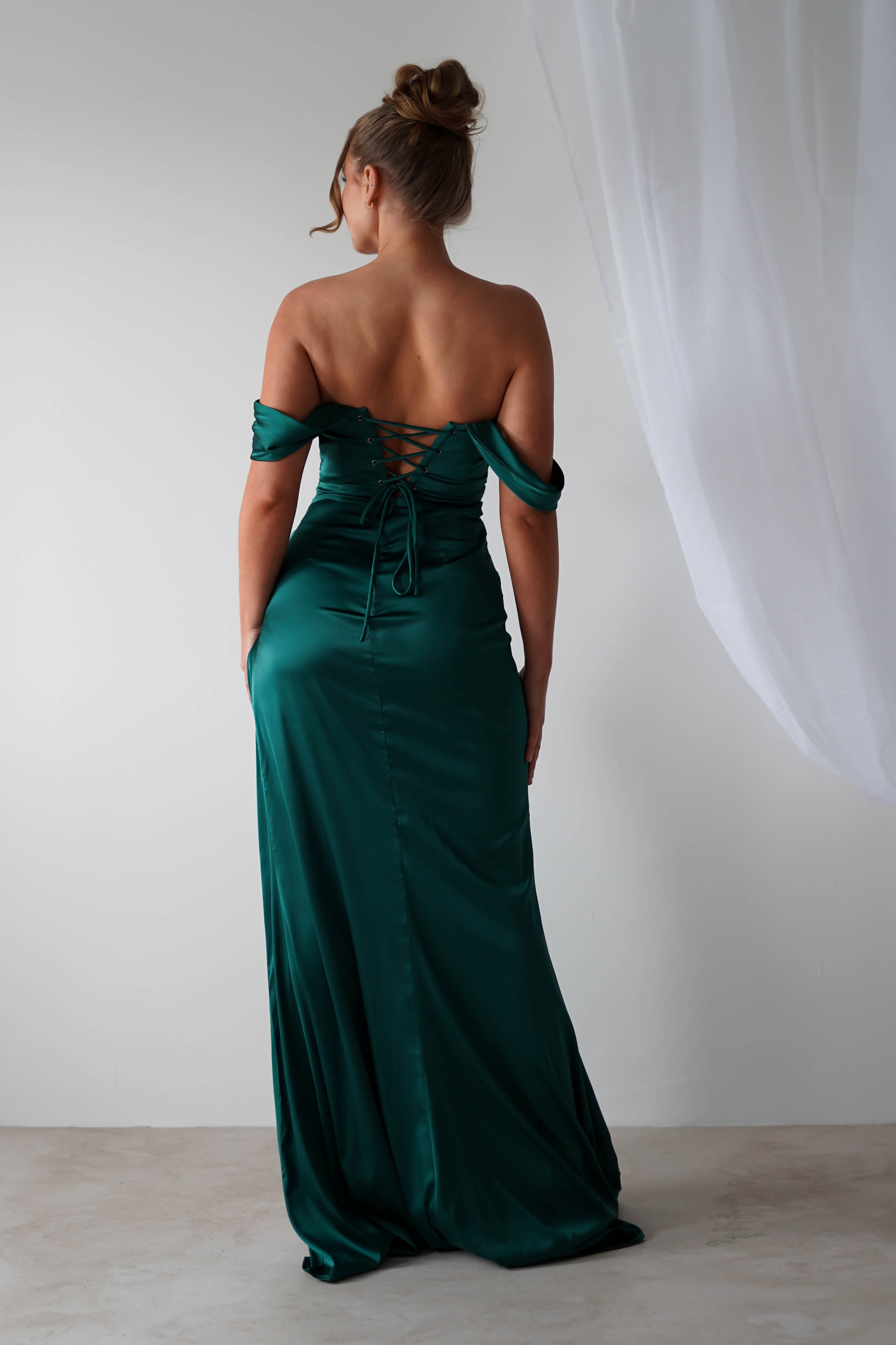 Woman standing with her back to the camera, wearing a long, off-the-shoulder gown in dark green satin material with lace-up detailing, in a minimalistic room with white walls and sheer curtains—an elegant choice for bridesmaids.