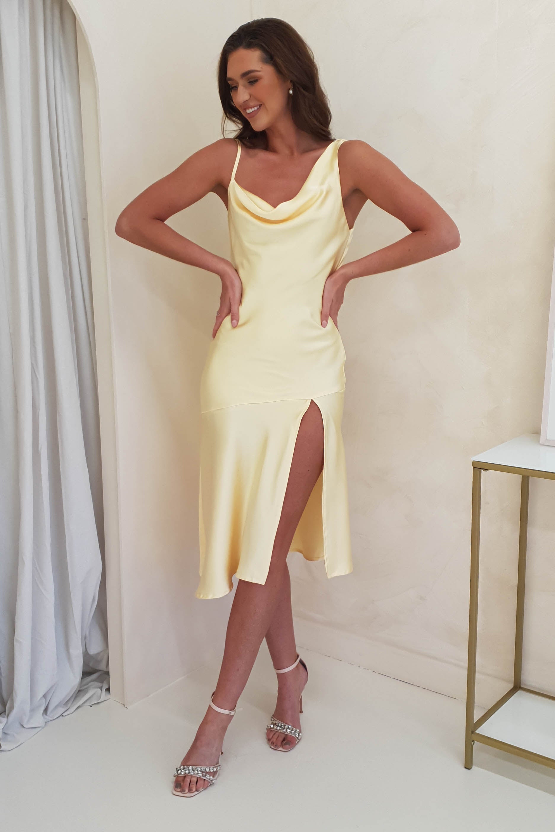 A woman in a sleeveless, light yellow, satin midi dress with a cowl neckline and open-back detail stands indoors, smiling with her hands on her hips. She wears strappy heels and has long wavy hair.