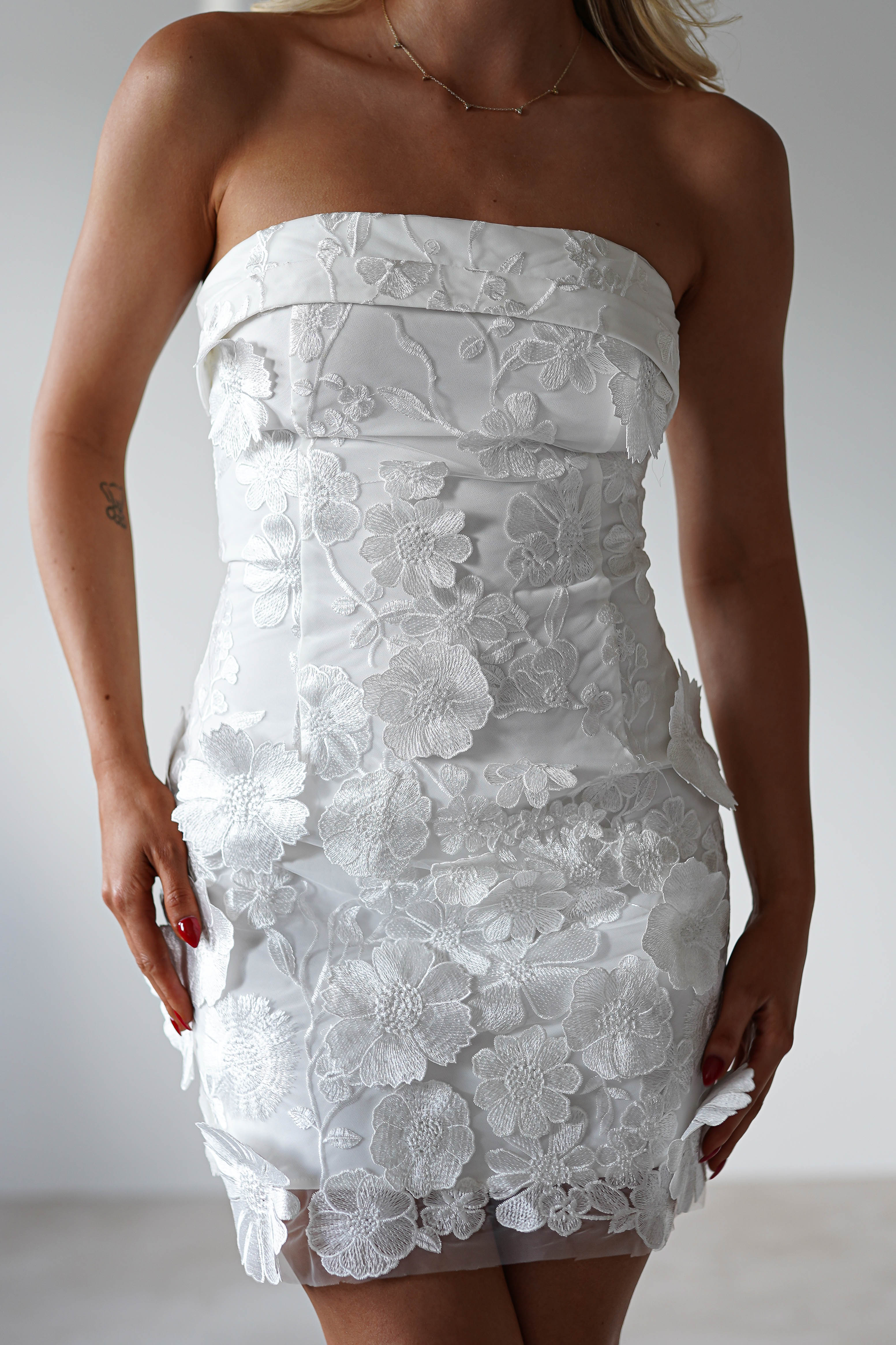 A woman is wearing a strapless mini dress adorned with floral lace patterns, crafted from exquisite floral 3D fabric. The fitted design highlights intricate flower details. Her red nails pop against her elegant look, and her hair cascades over one shoulder. The background is neutral and modern.