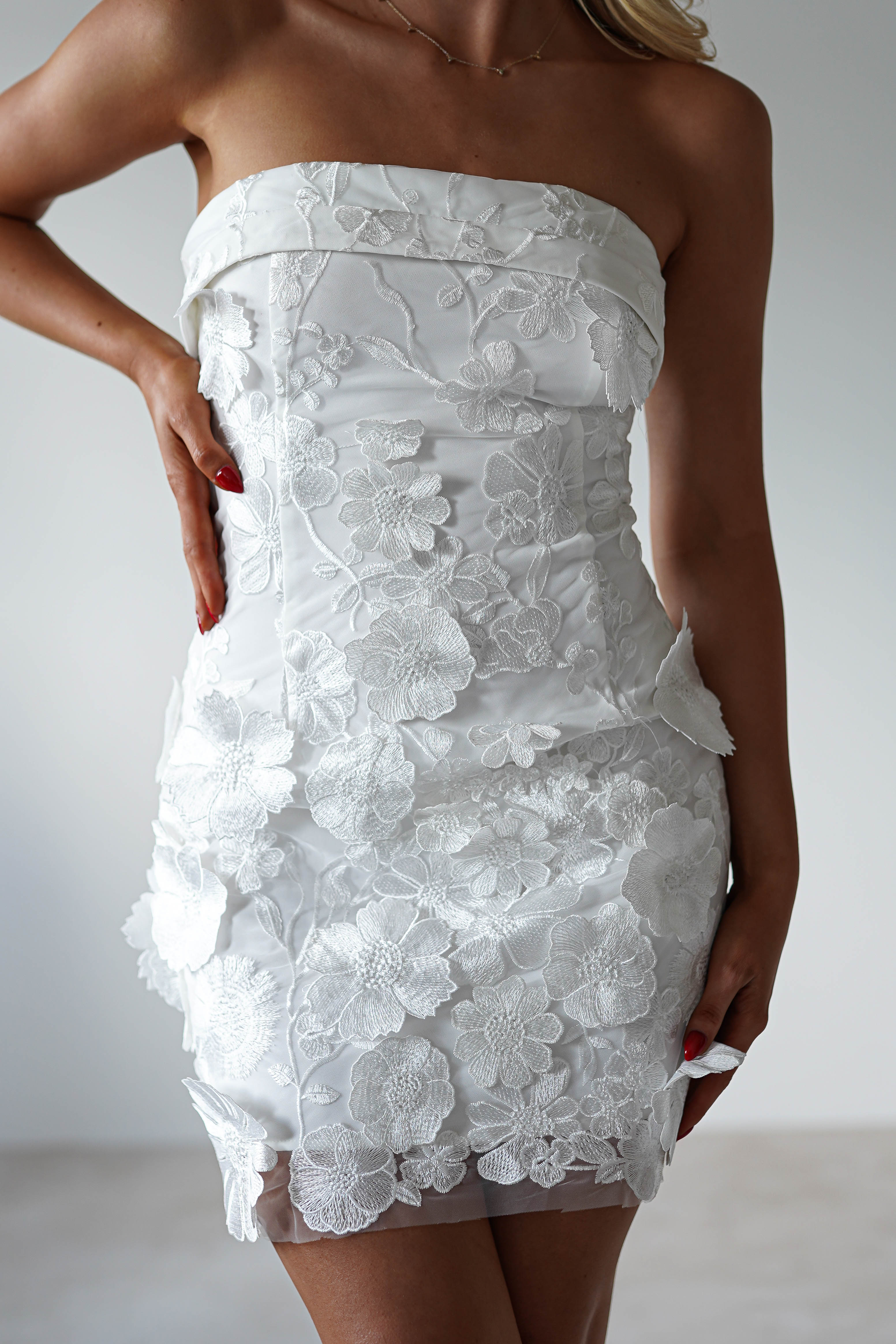 A person is wearing an elegant white, strapless mini dress adorned with lace floral patterns. Their hand rests on their hip, highlighting the dresss fitted design. Perfect wedding guest attire against a plain, light background.