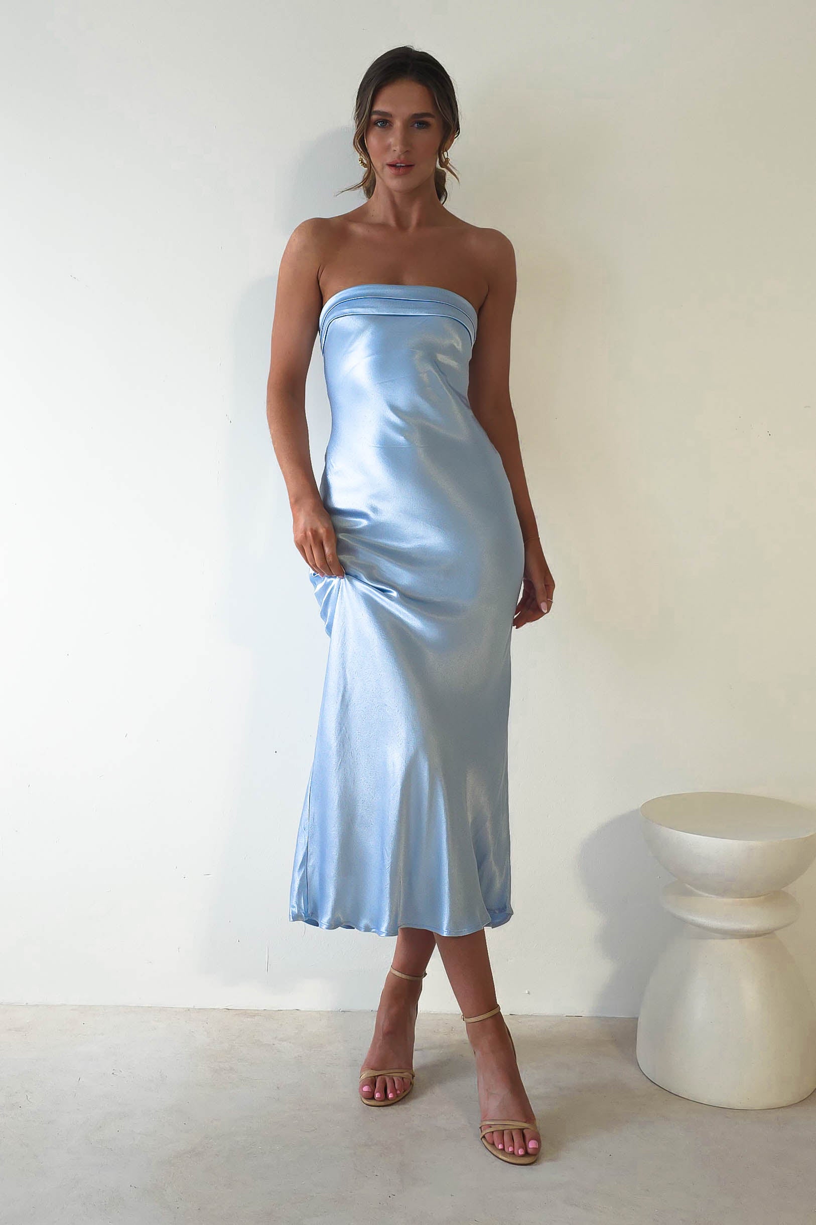 A woman stands against a plain white wall, dressed in a strapless midi dress of light blue satin that drapes below the knees. Her hair is tied back as she wears high-heeled sandals. Ideal for wedding guests, this ensemble pairs perfectly with the white, round pedestal on her right.