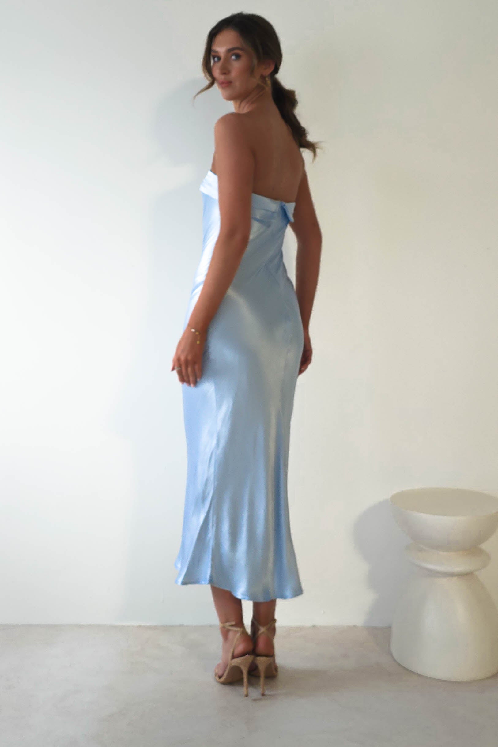 A woman stands in a simple, well-lit room, wearing a strapless midi dress in light blue satin and beige high heels. Her hair is tied back as she turns slightly, glancing back. A small, round, white table is beside her—perfect attire for stylish wedding guests embracing elegance.