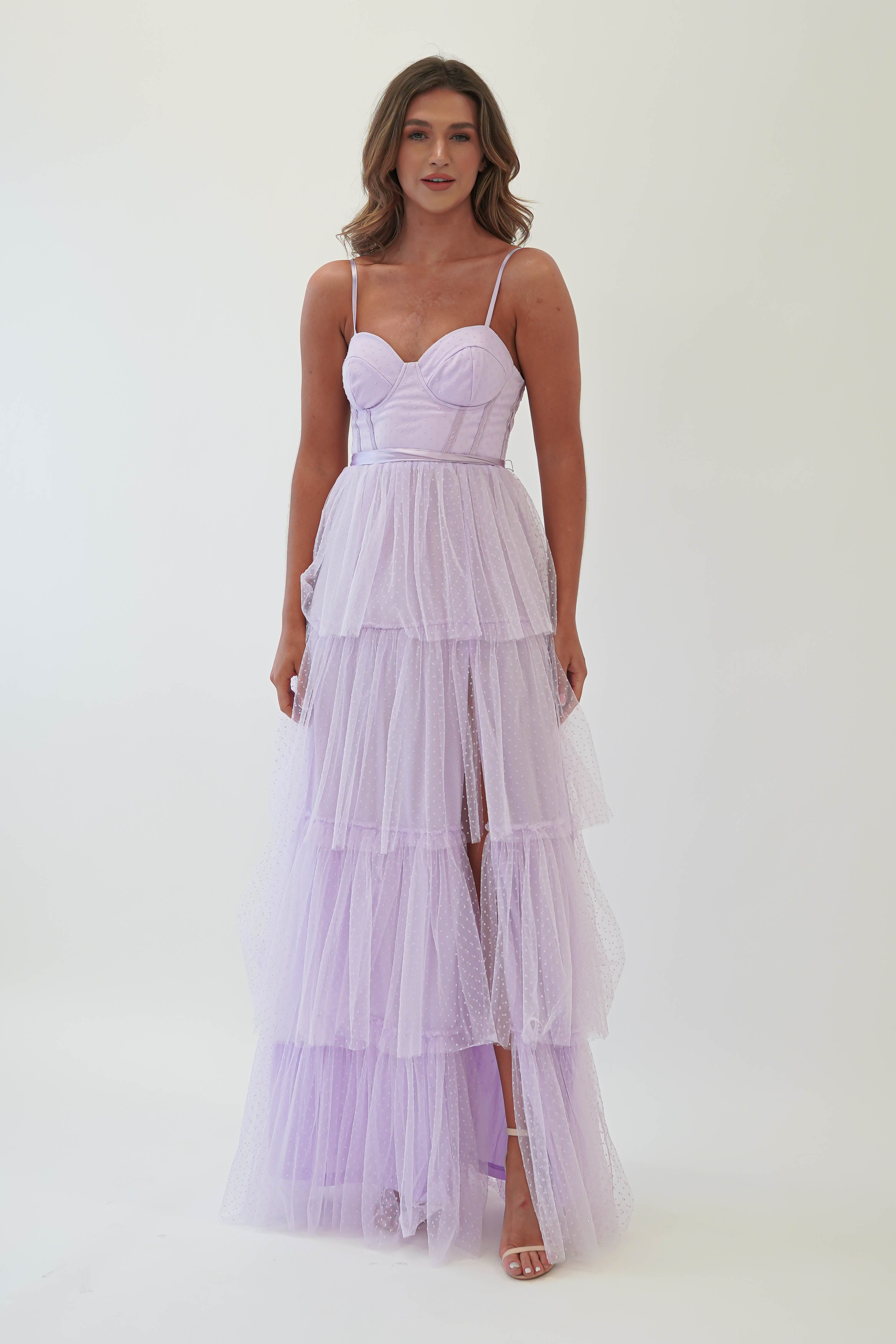 A person wearing a lavender tulle maxi dress with spaghetti straps stands against a plain white background. Perfect for formal events, the gown features a fitted bodice with a sweetheart neckline, flowing elegantly into a long, tiered skirt.