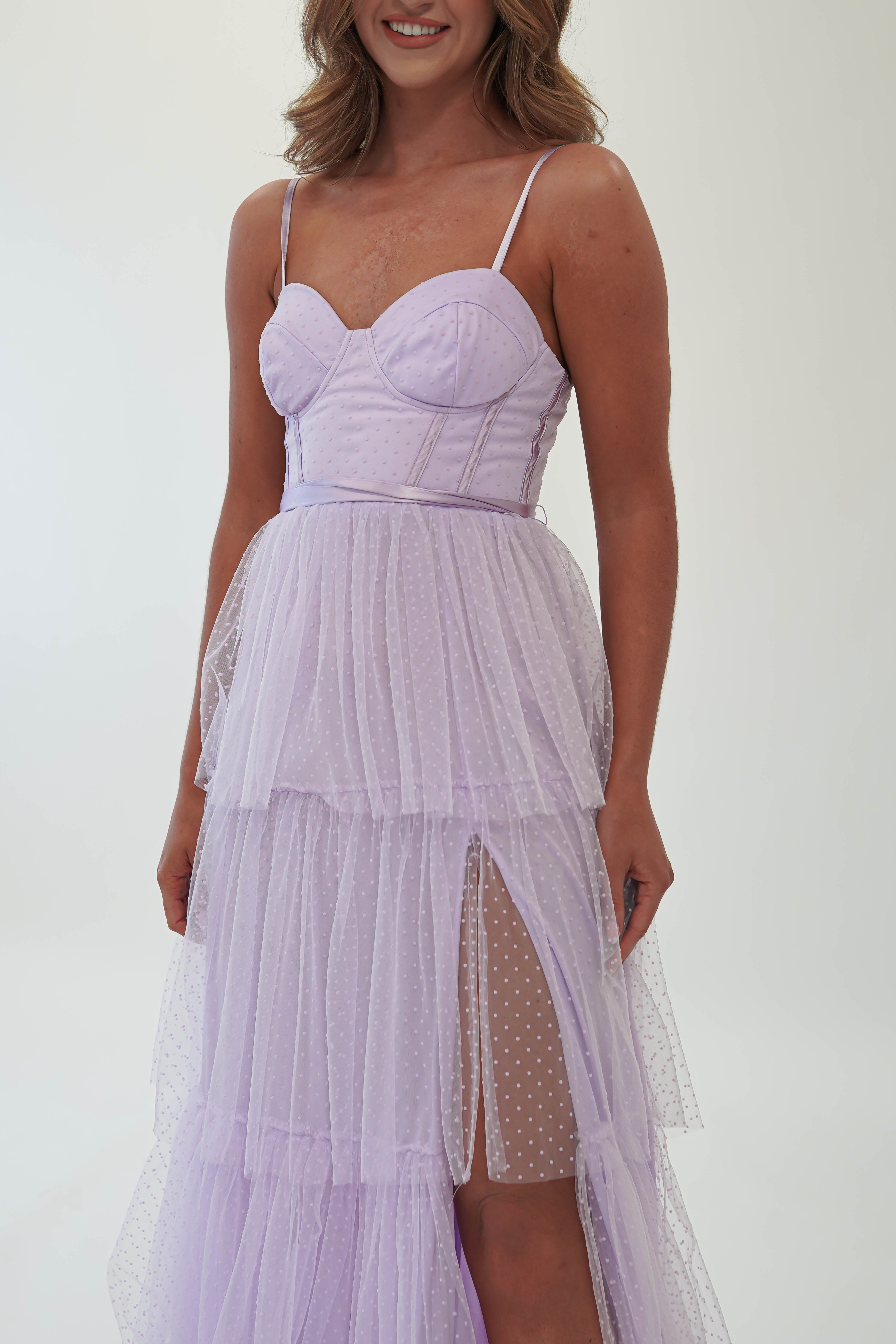 A woman wearing a light purple, sleeveless spaghetti strap dress with a fitted bodice and layered, flowing skirt featuring a thigh-high slit. Perfect as a wedding guest outfit, the fabric has a subtle polka dot pattern. She stands against a plain white background.