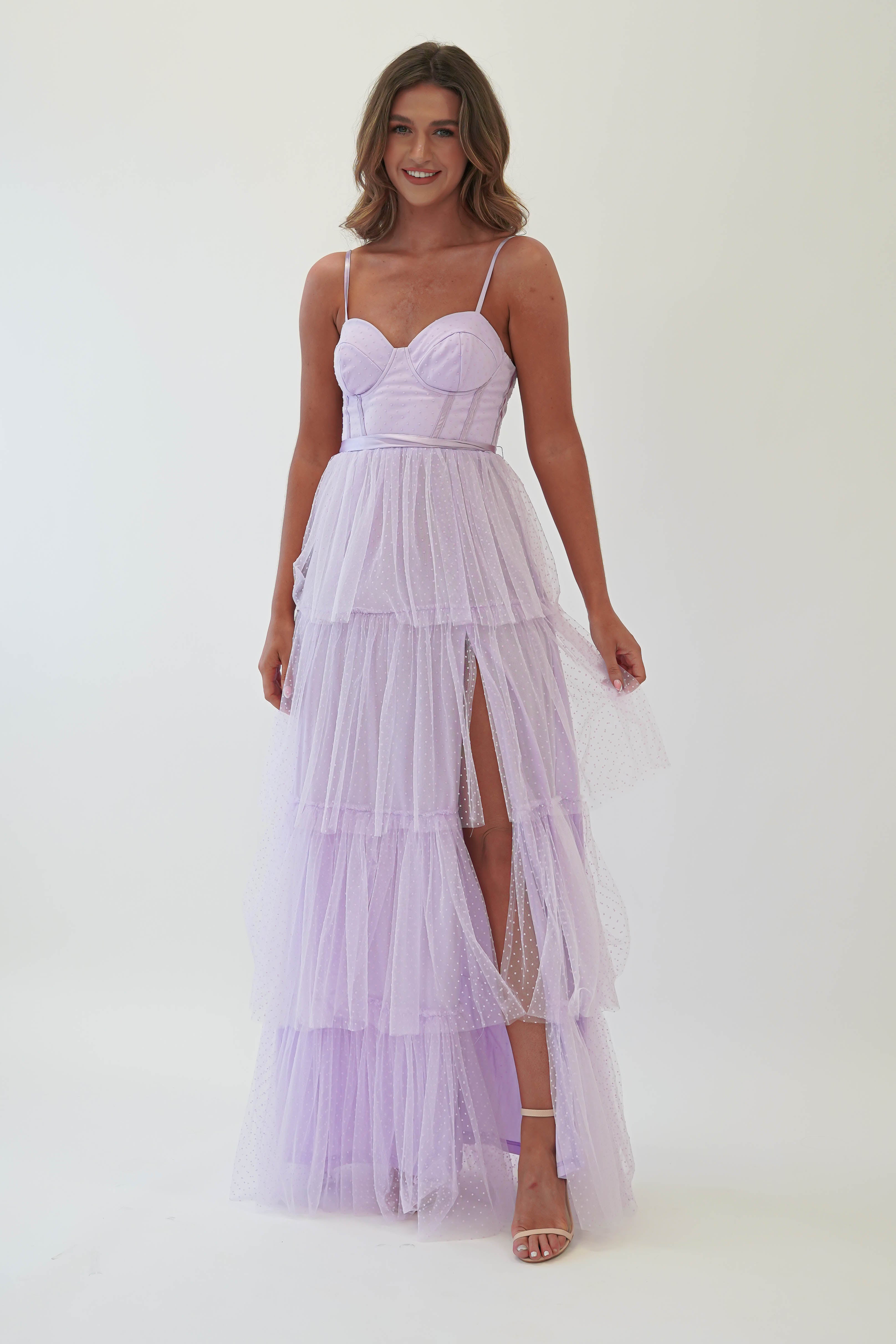 A woman in a lavender tulle maxi dress with a sweetheart neckline and spaghetti straps stands against a plain background. Perfect for formal events, the gown features a high slit. She is wearing high heels and smiling slightly, her left hand gracefully holding the dress.