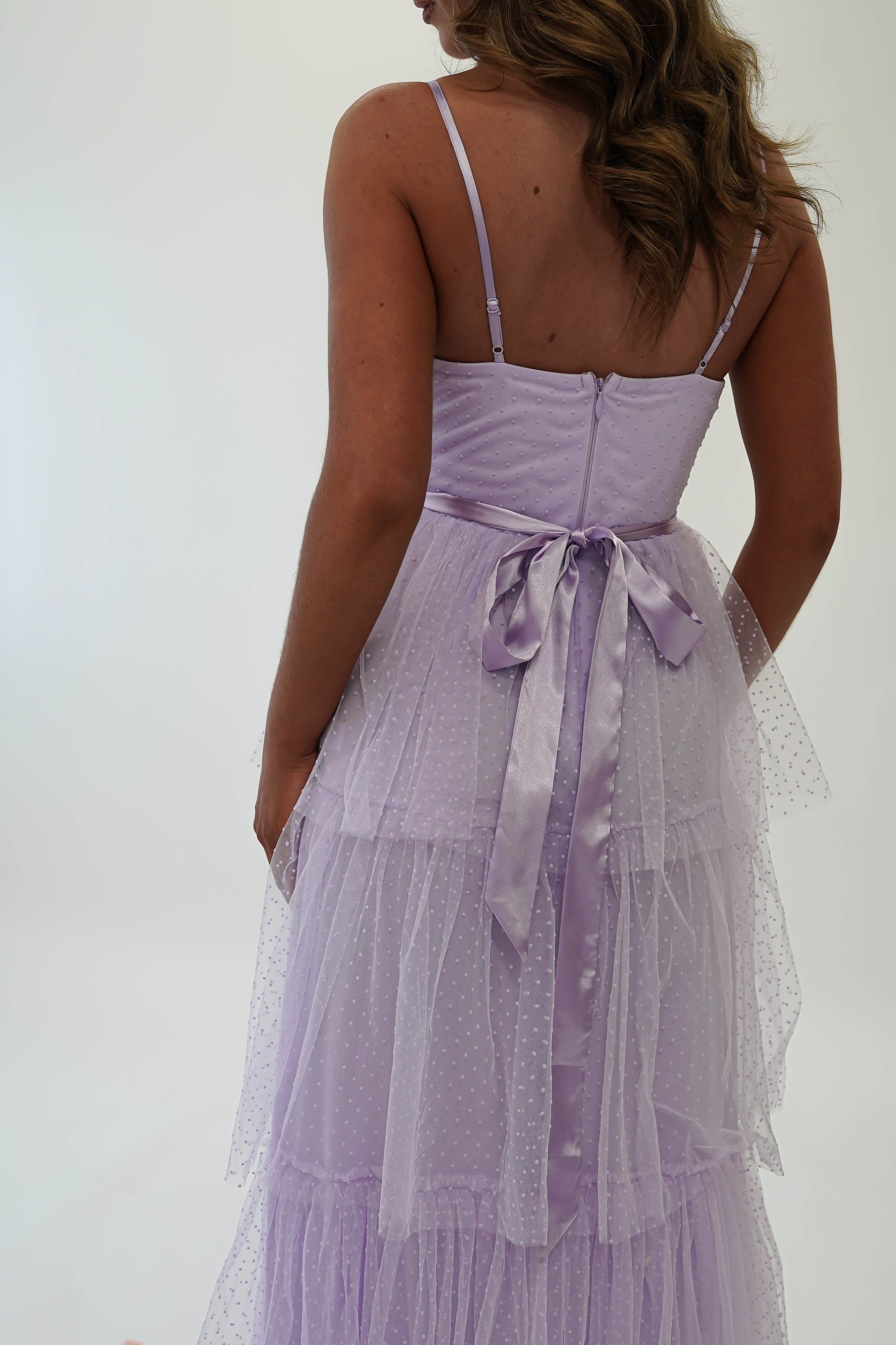 Rear view of a woman in a light purple, polka-dotted tulle maxi dress with a sheer, tiered skirt and a ribbon bow at the waist. Perfect for formal events or as a wedding guest outfit, this dress has spaghetti straps and a back zipper against a plain white background.