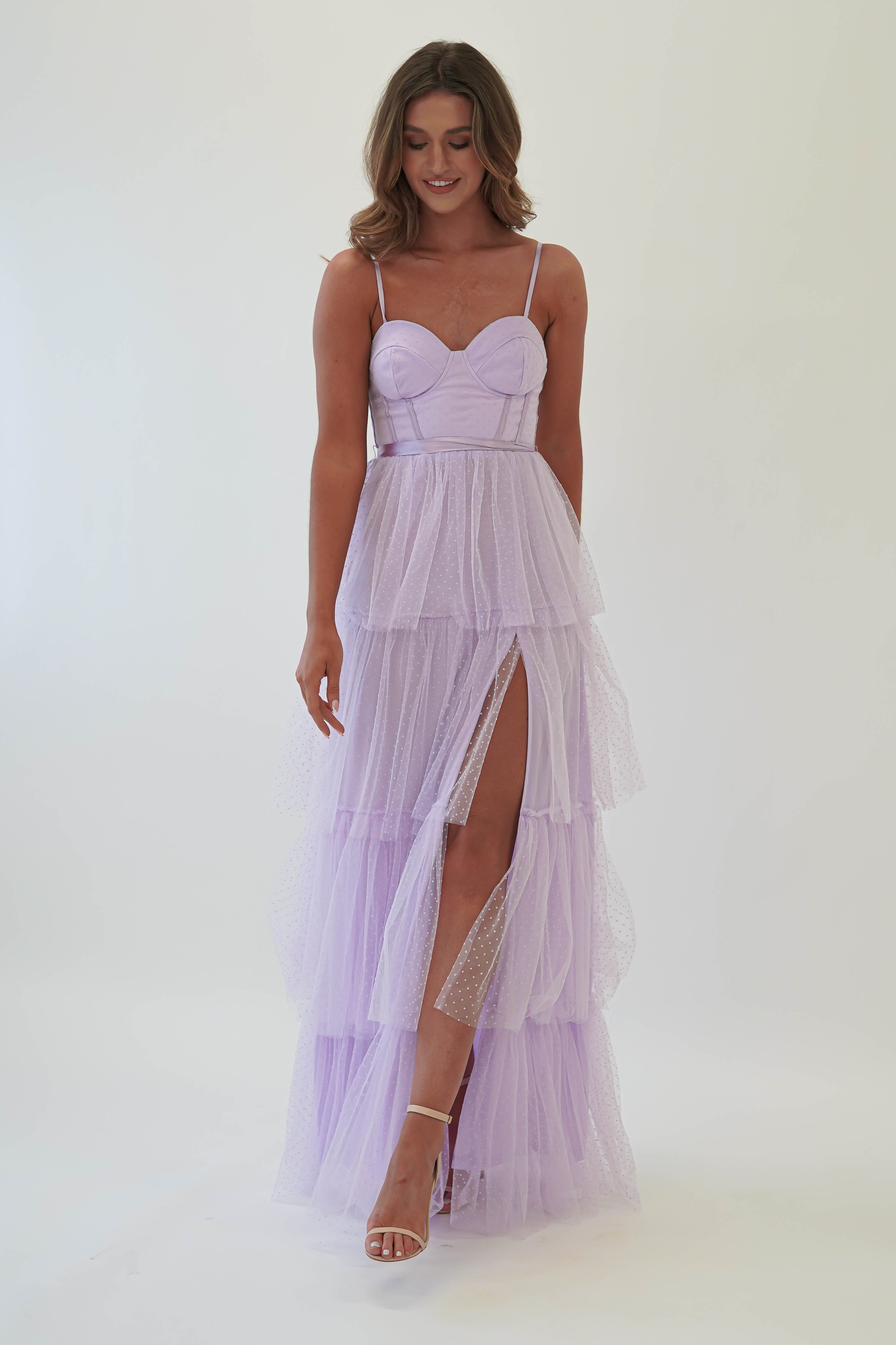 A woman in a lilac tulle maxi dress with spaghetti straps and a sweetheart neckline stands against a white background. Perfect for formal events or as a wedding guest, the dress features a high slit and sheer layers. She completes the look with white high heels and shoulder-length hair.