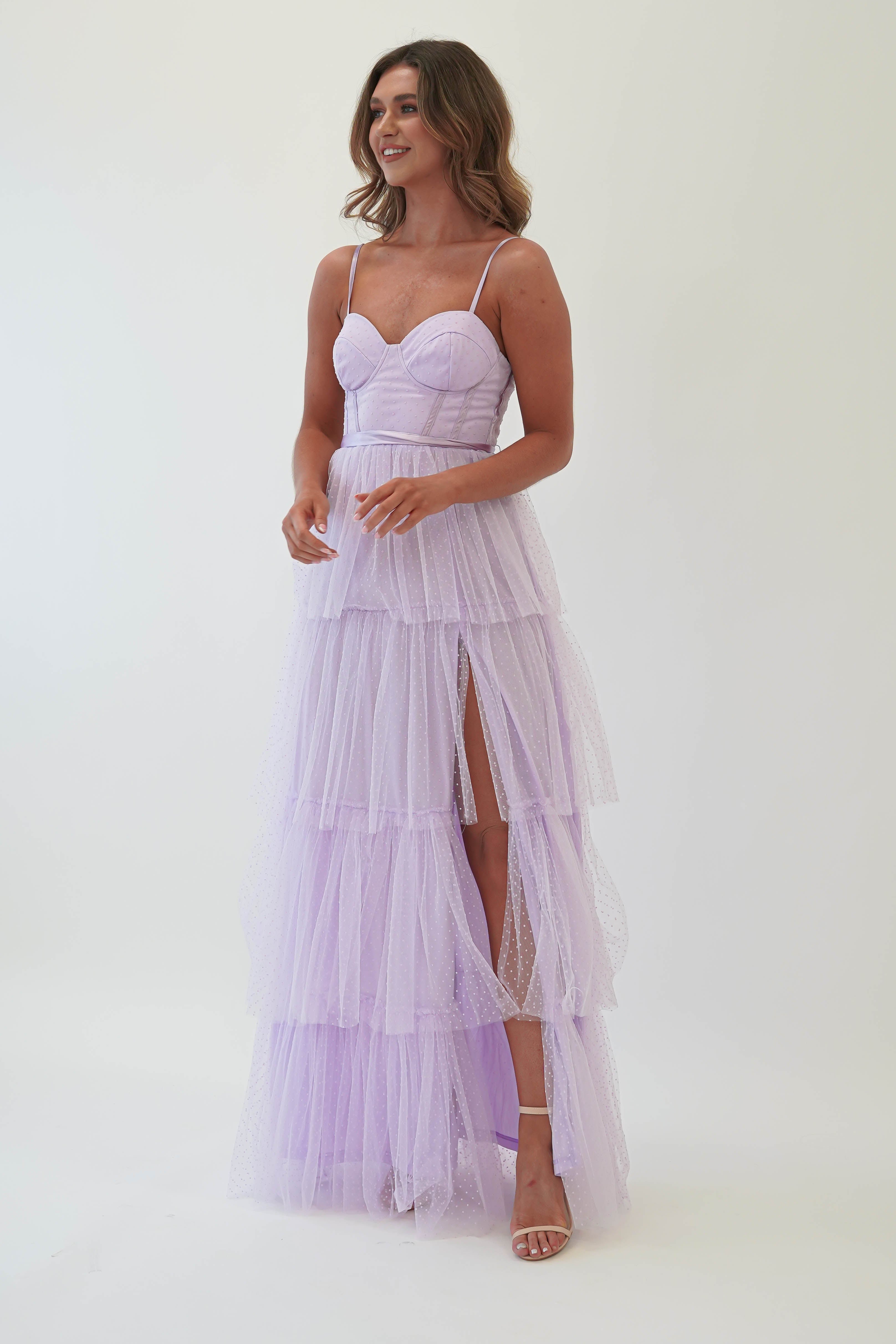 A woman is wearing a lavender tulle maxi dress with spaghetti straps and a thigh-high slit, perfect for formal events. She has long, wavy hair and is posing against a plain white background, exuding elegance suitable for a wedding guest.