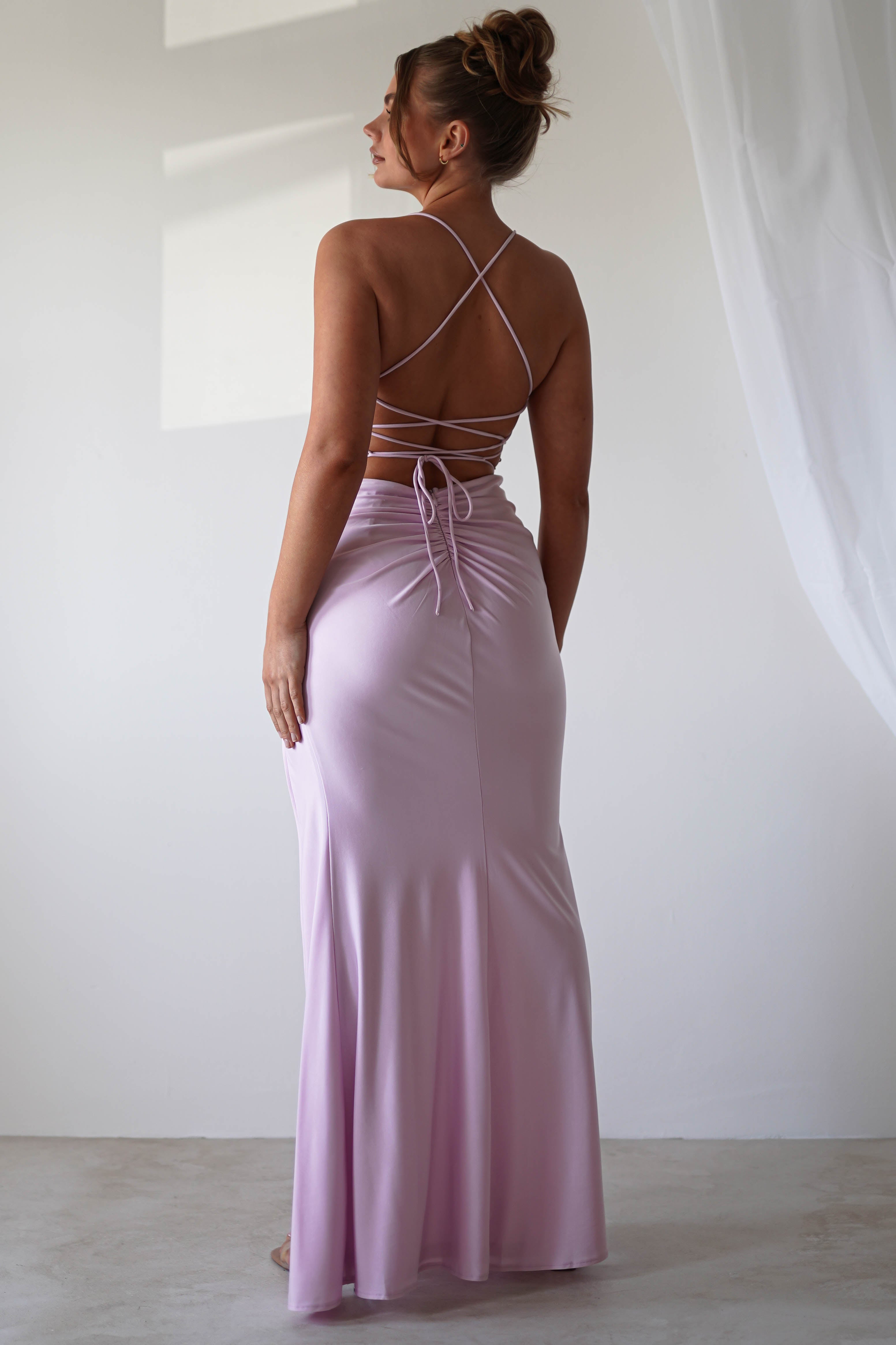 Woman facing away, wearing a long, backless lavender gown with crisscross straps and sequin detail accentuating the ruched waist. Her hair is styled in an updo. The setting is indoors with soft lighting and sheer curtains in the background, perfect for formal events.